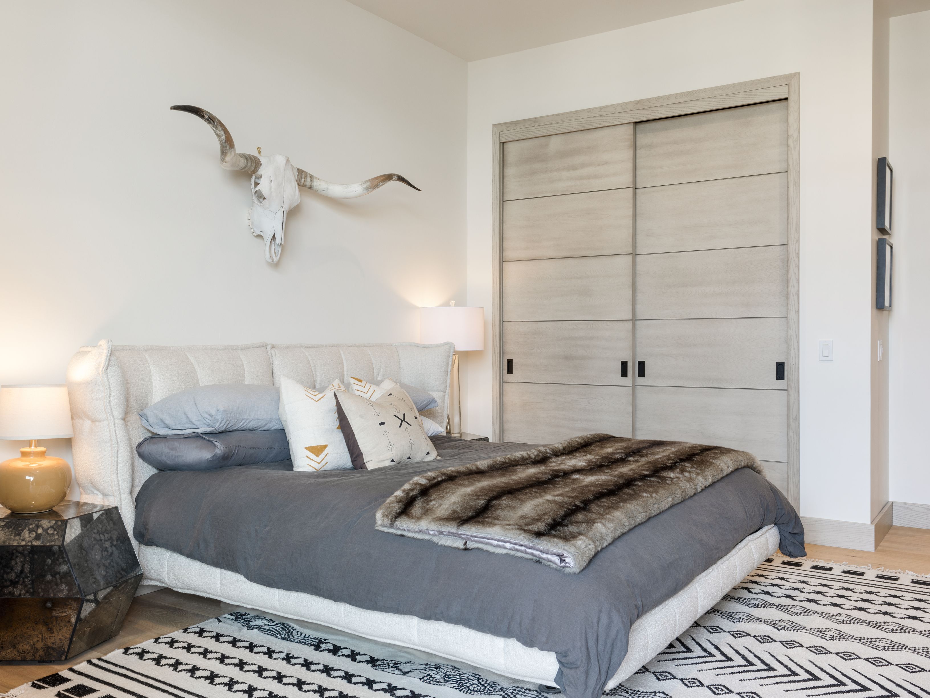 This is a well-lit primary bedroom featuring a large bed with a tufted headboard, neutral-toned bedding, and decorative pillows. A faux fur throw is draped across the bed. A steer skull is mounted on the wall above the bed, adding a rustic touch. Sliding closet doors are visible on the right side of the image, and a geometric patterned rug is on the floor.