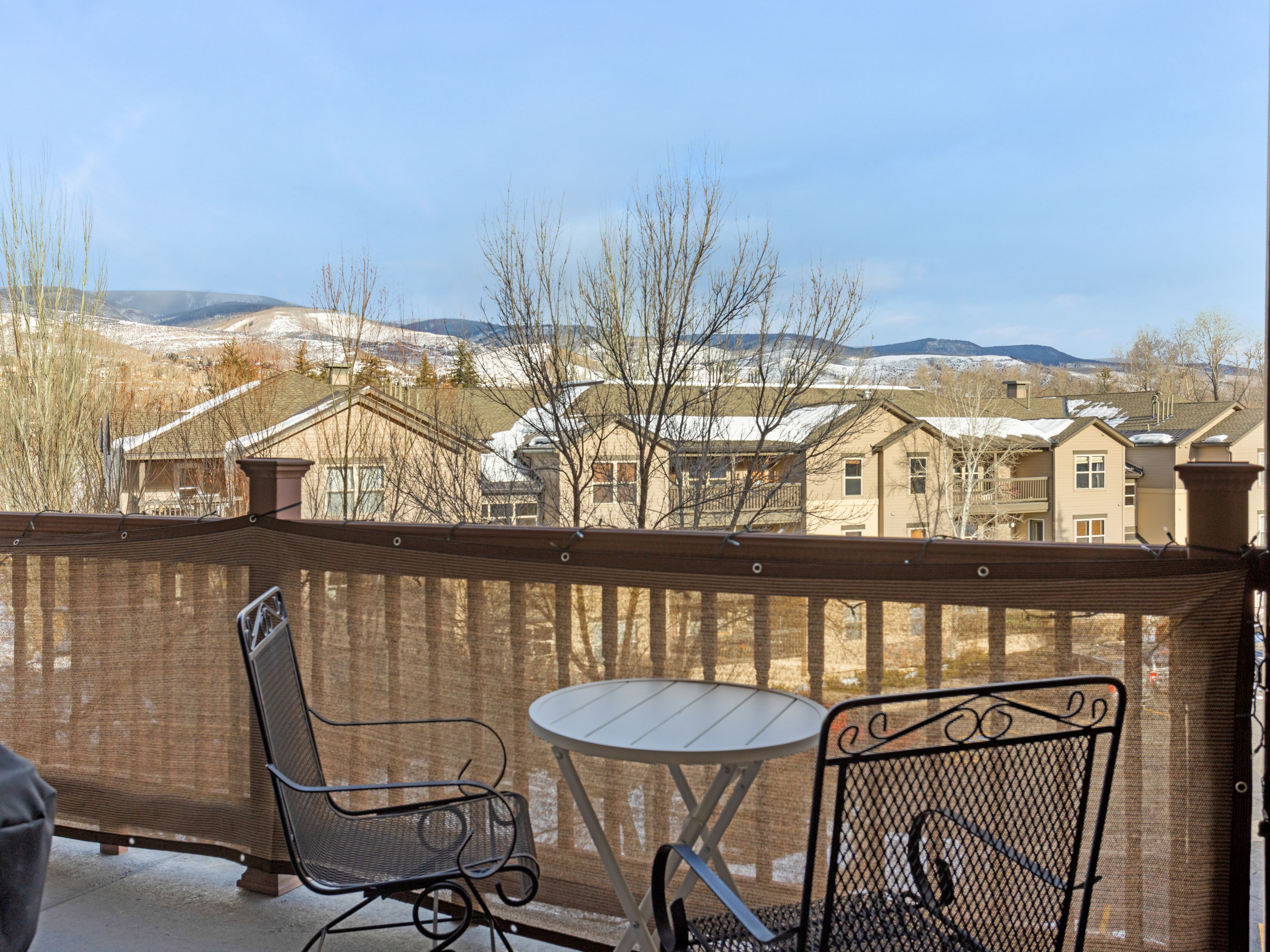 This image showcases a cozy balcony setting with a small round table and two wrought iron chairs, creating an inviting outdoor space. A privacy screen provides seclusion while still allowing a view of the surrounding neighborhood and distant snow-capped mountains. The scene suggests a relaxing spot for enjoying the scenery and fresh air.