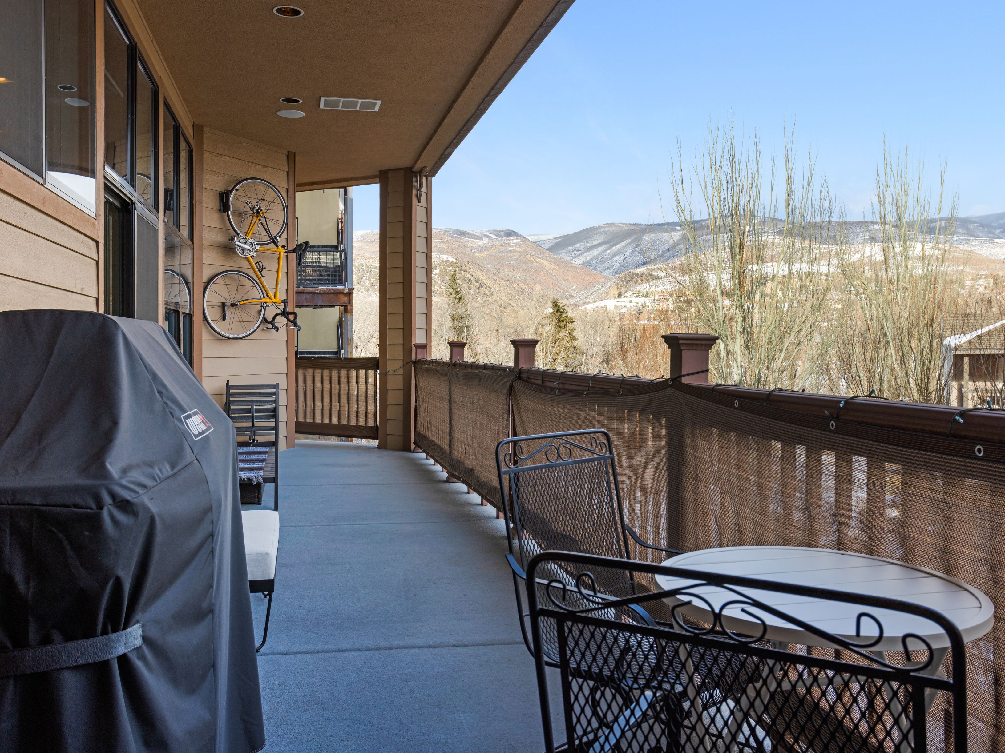 This image showcases a covered balcony with outdoor seating and a view of distant mountains. The balcony features a grill, a small round table with metal chairs, and a privacy screen along the railing. A yellow bicycle is mounted on the wall, adding a touch of personality to the outdoor space.