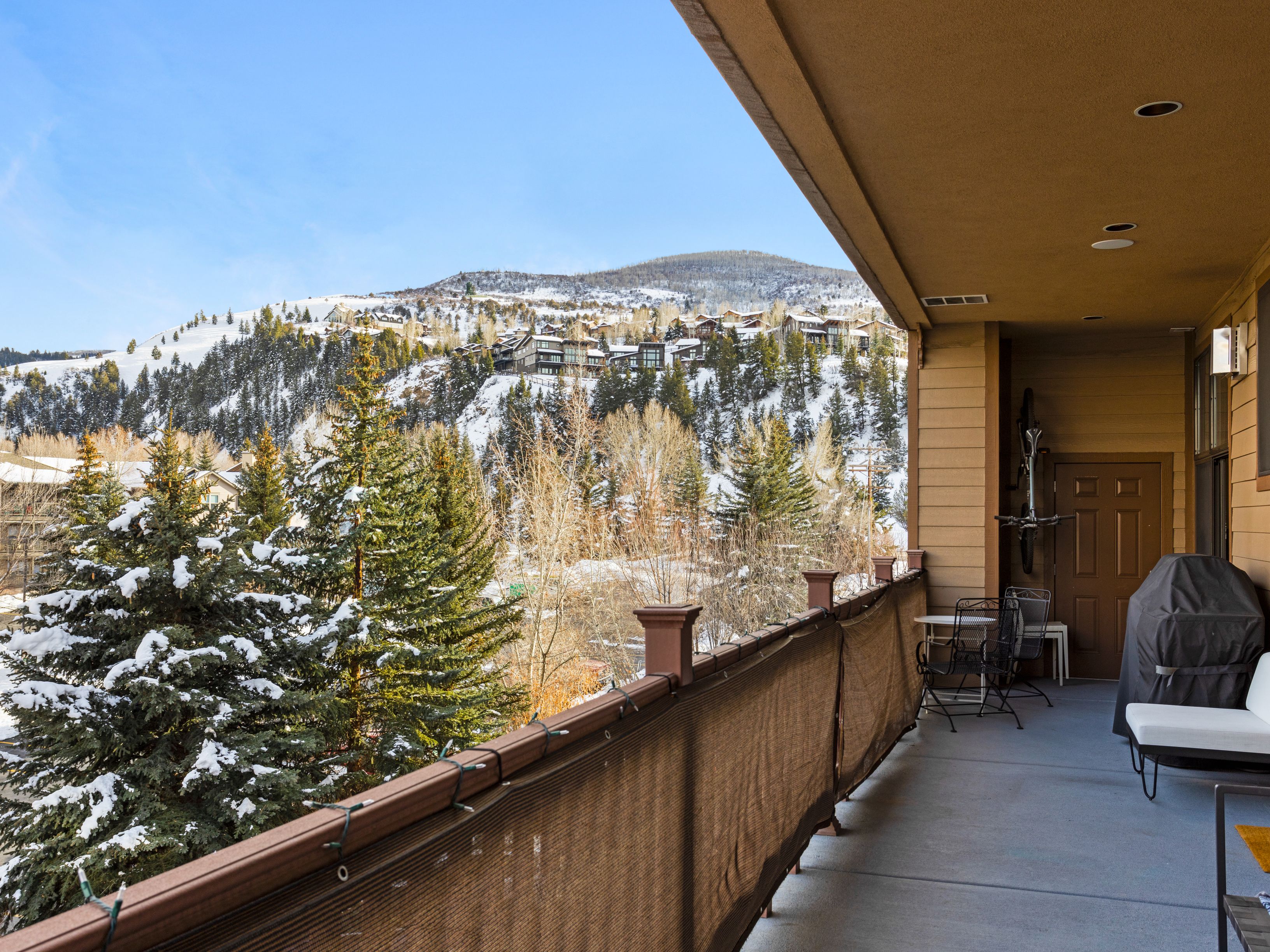 This image showcases a spacious balcony with a view of a snow-covered landscape. The balcony features outdoor furniture, including a table, chairs, and a grill, suggesting a comfortable space for relaxation and entertainment. The view includes snow-covered trees and a hillside with houses, adding to the appeal of the outdoor living space.