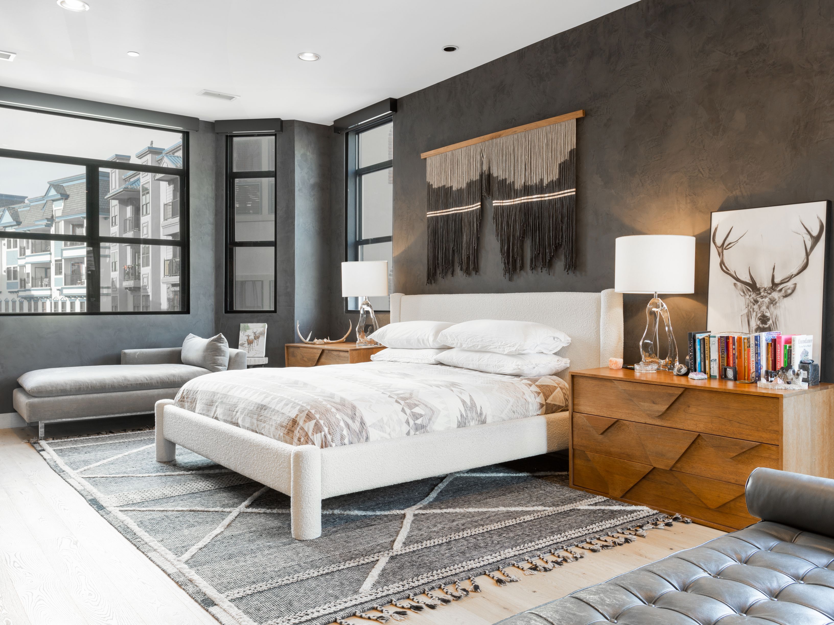 This is a stylish primary bedroom featuring a large bed with a textured white frame and neutral bedding. The room has a modern aesthetic with dark gray walls, large windows providing natural light, and wooden nightstands. A decorative wall hanging and a deer print add character to the space, while a gray rug anchors the room.