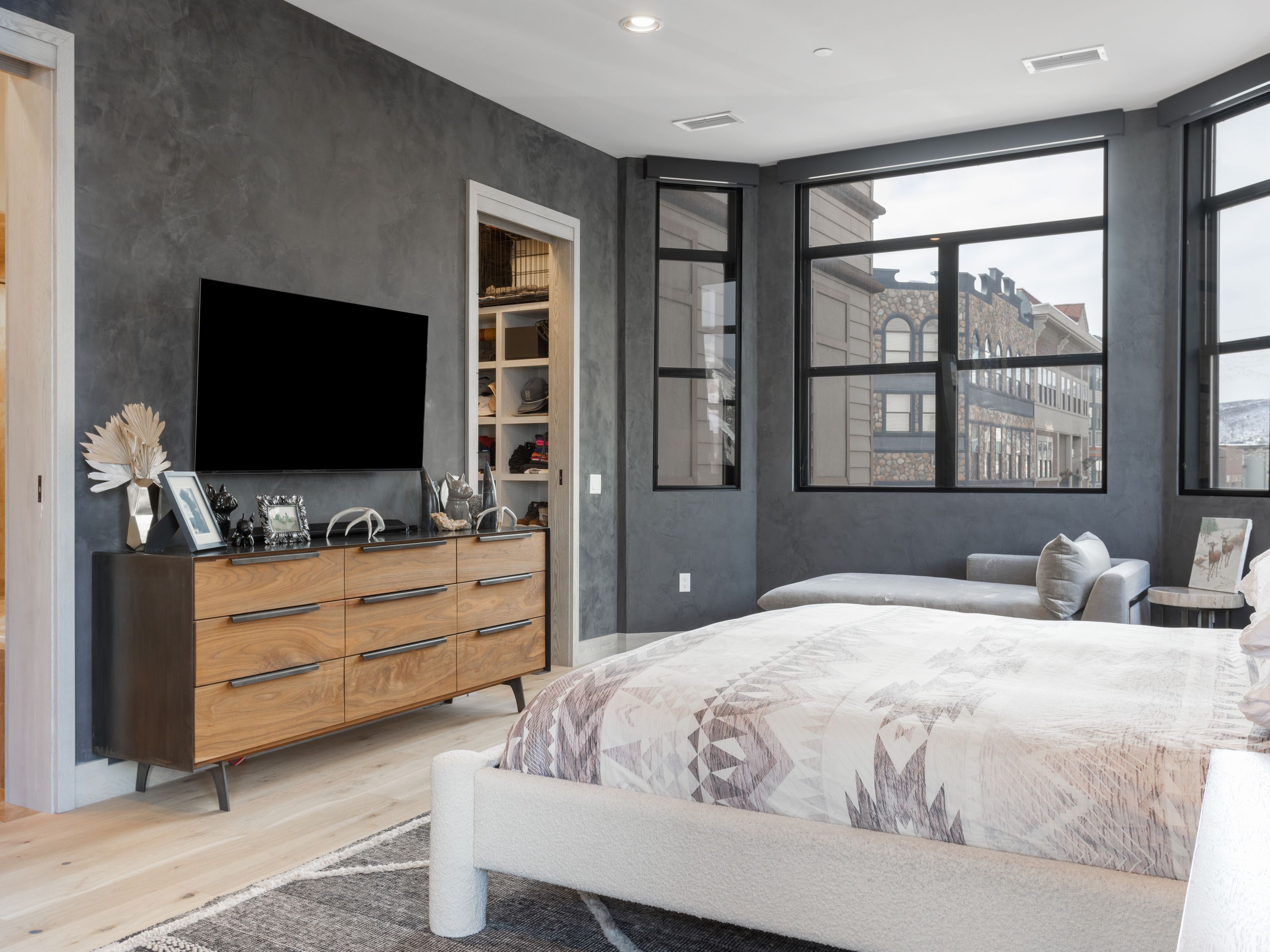 This is a primary bedroom featuring a modern design with a gray accent wall and a large window offering an exterior view. The room includes a wooden dresser with a television mounted above it, a walk-in closet, and a bed with a patterned comforter. The overall impression is luxurious and comfortable.