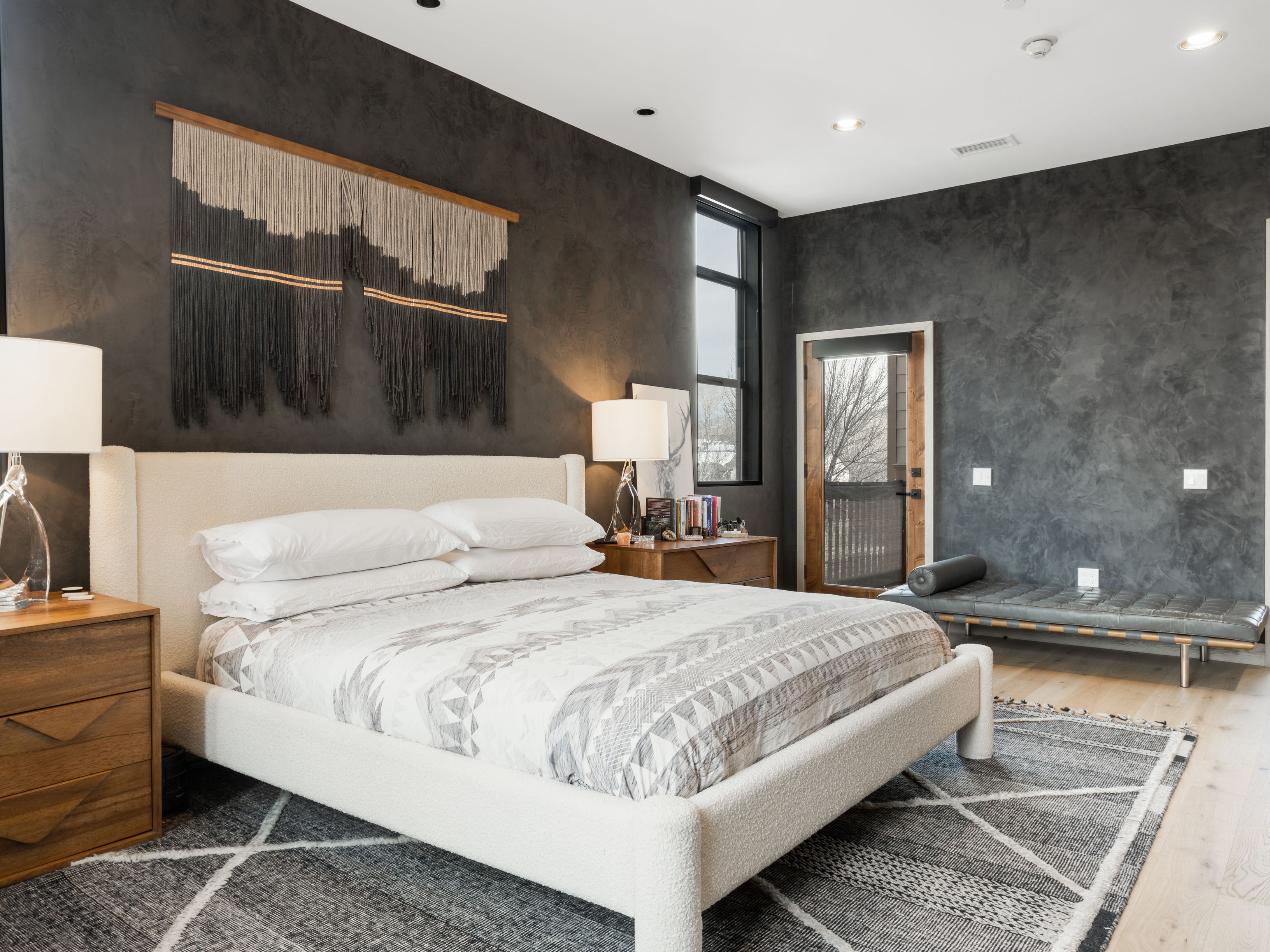 This is a primary bedroom featuring a large bed with a white frame and patterned bedding, flanked by wooden nightstands with lamps. The walls are a dark, textured gray, and a woven wall hanging adds visual interest above the bed. A gray rug anchors the space, and a bench sits near a door leading outside, creating a modern and inviting atmosphere.