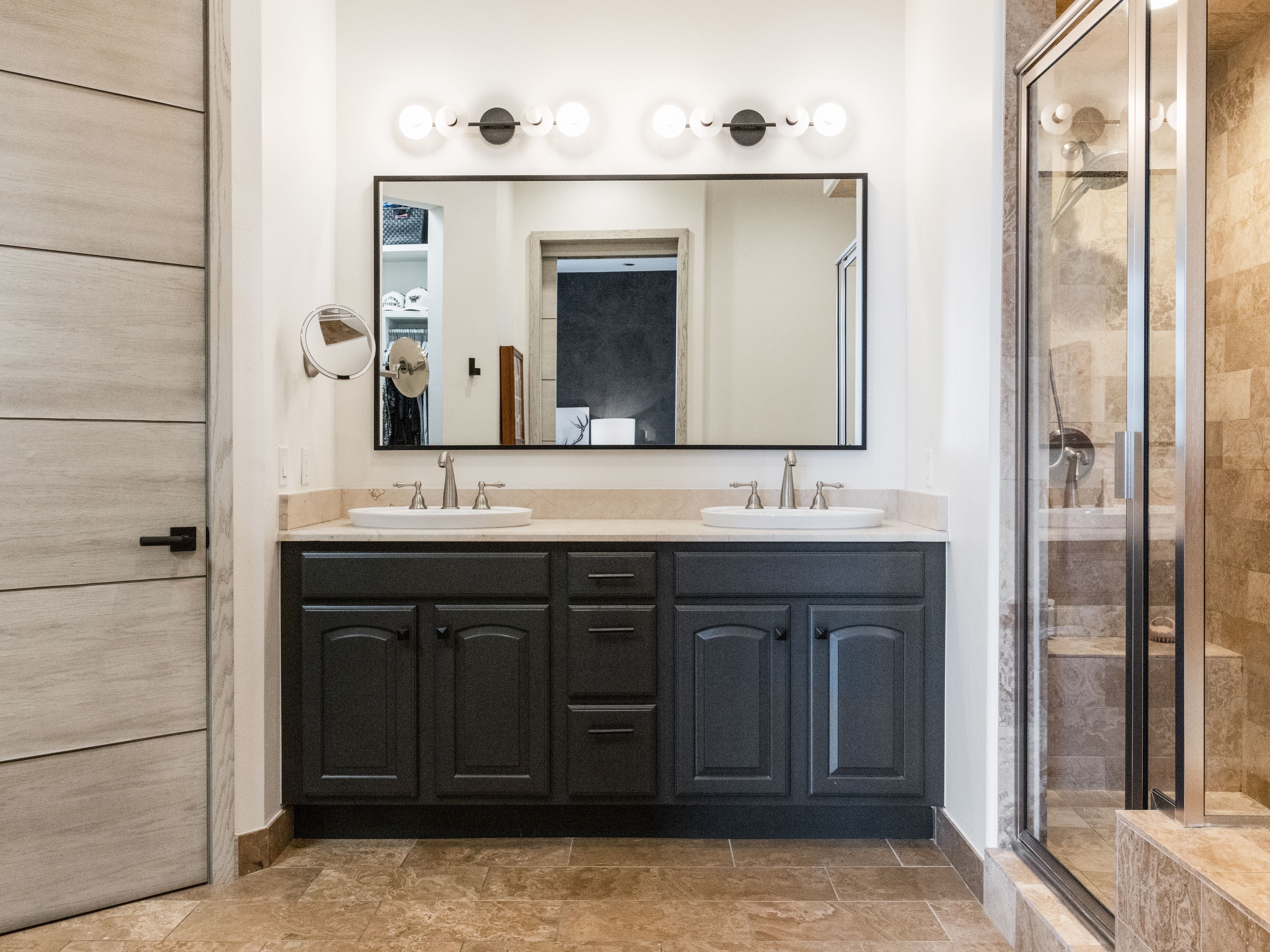 This is a well-lit primary bathroom featuring a double vanity with dark cabinetry and light countertops. A large mirror spans the width of the vanity, reflecting the space and enhancing the sense of openness. A glass-enclosed shower is visible to the right, suggesting a modern and luxurious bathing experience. The perspective is from the doorway, showcasing the entire vanity area and part of the shower.