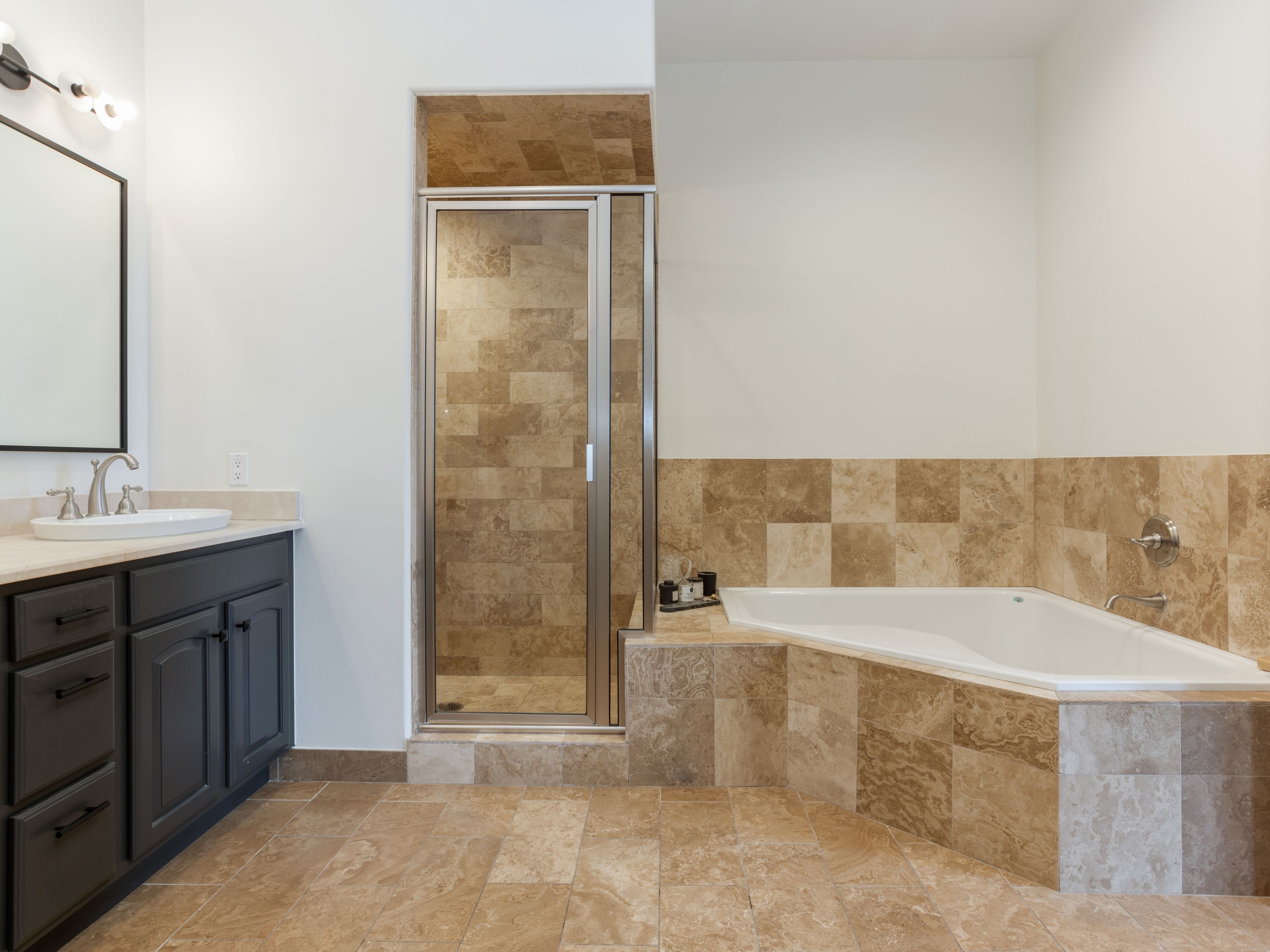 This is a primary bathroom featuring a dark wood vanity with a light countertop and a vessel sink. A large mirror hangs above the sink, illuminated by sconce lighting. The bathroom also includes a glass-enclosed shower and a soaking tub, both surrounded by earth-toned tile, creating a cohesive and luxurious spa-like atmosphere.