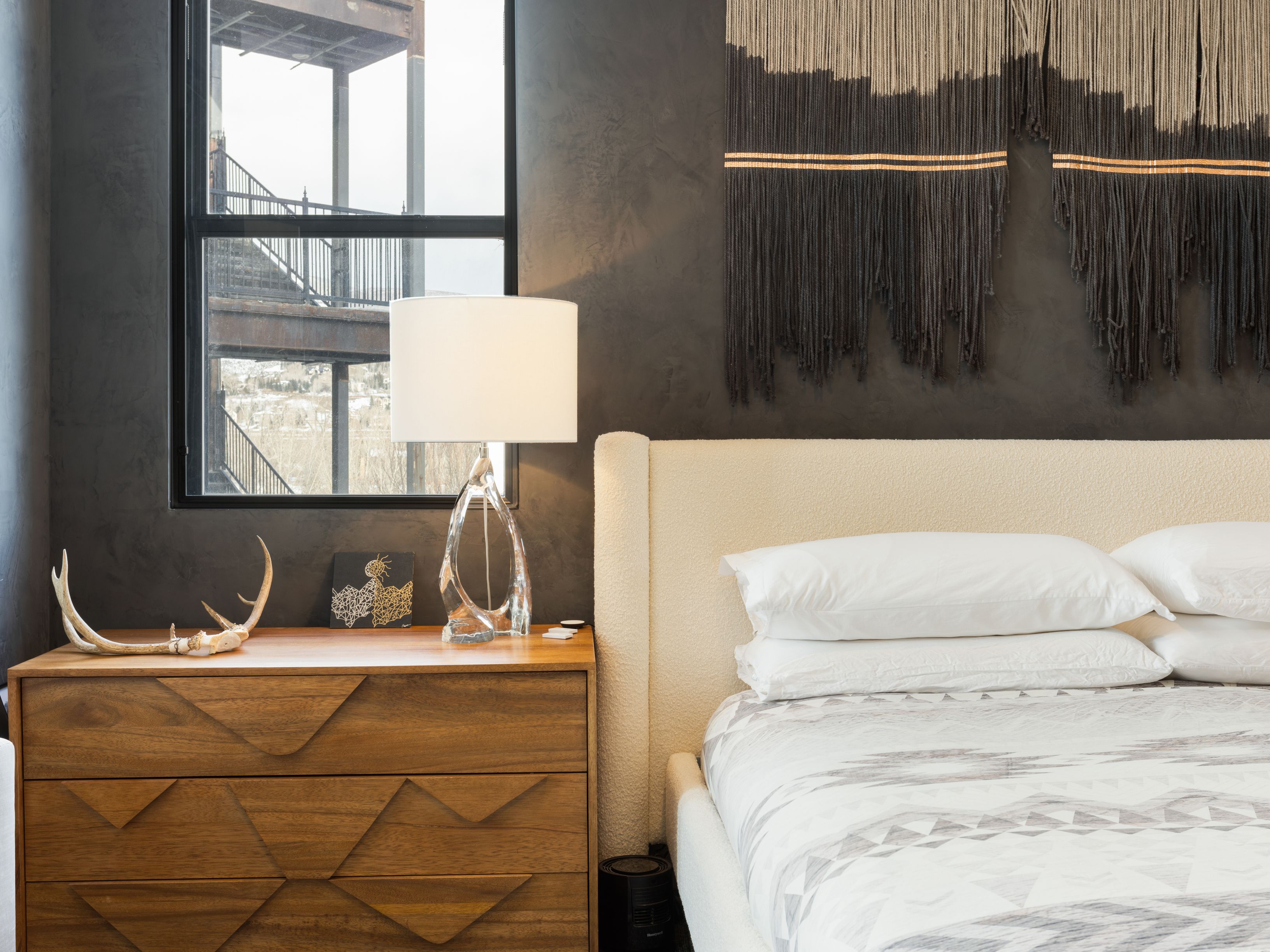 This is a stylish primary bedroom featuring a neutral color palette with a textured wall hanging above the bed. A wooden dresser with geometric detailing is positioned next to a window, displaying decorative items such as antlers and a modern lamp. The room exudes a modern and sophisticated ambiance.