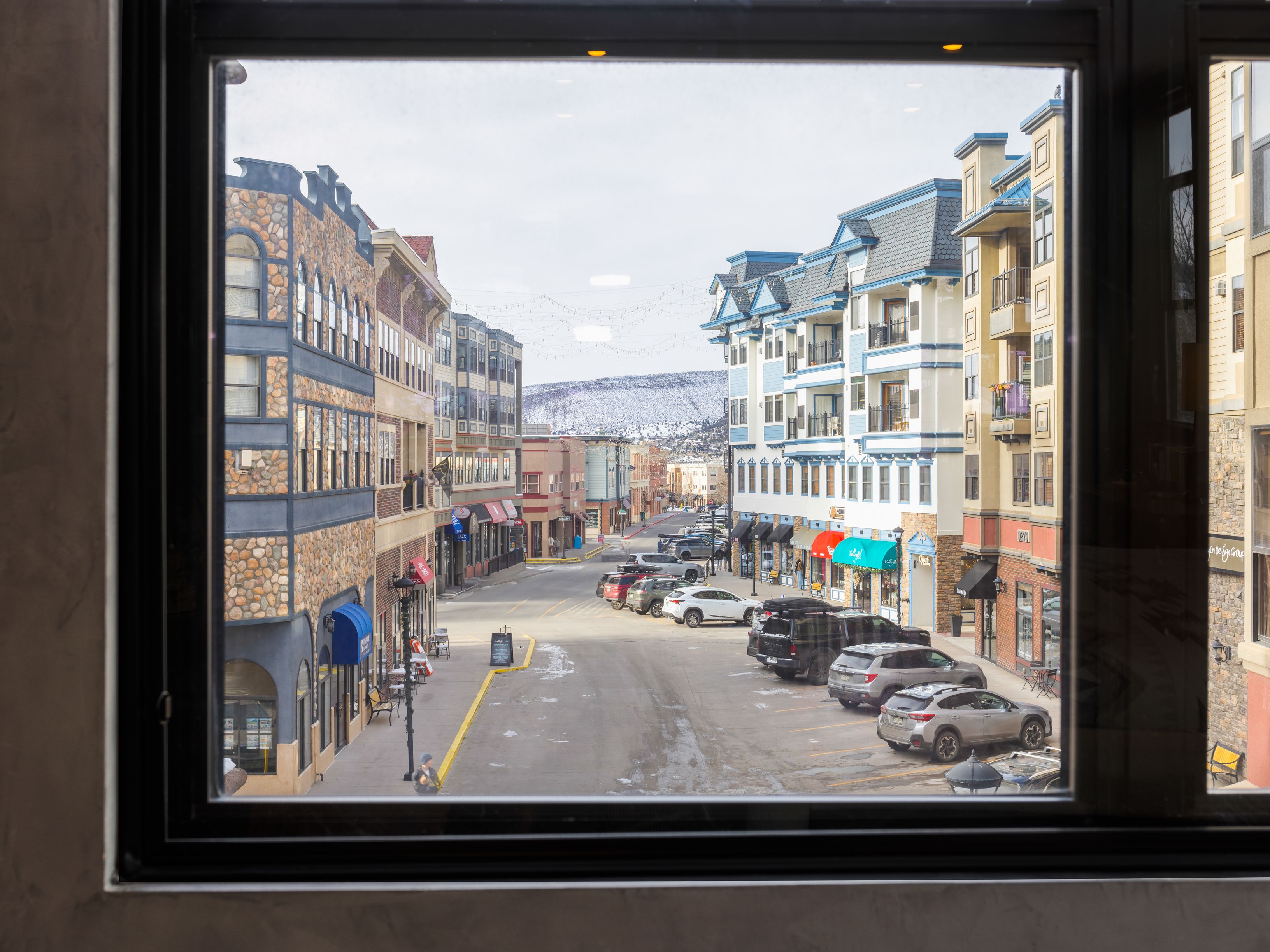 The image showcases a street view framed by a window, presenting a charming town with multi-story buildings exhibiting varied architectural styles, from stone facades to blue-trimmed structures. Cars are parked along the street, and the scene is set against a backdrop of distant, snow-capped mountains under a slightly overcast sky. The perspective is from inside, looking out onto the street.
