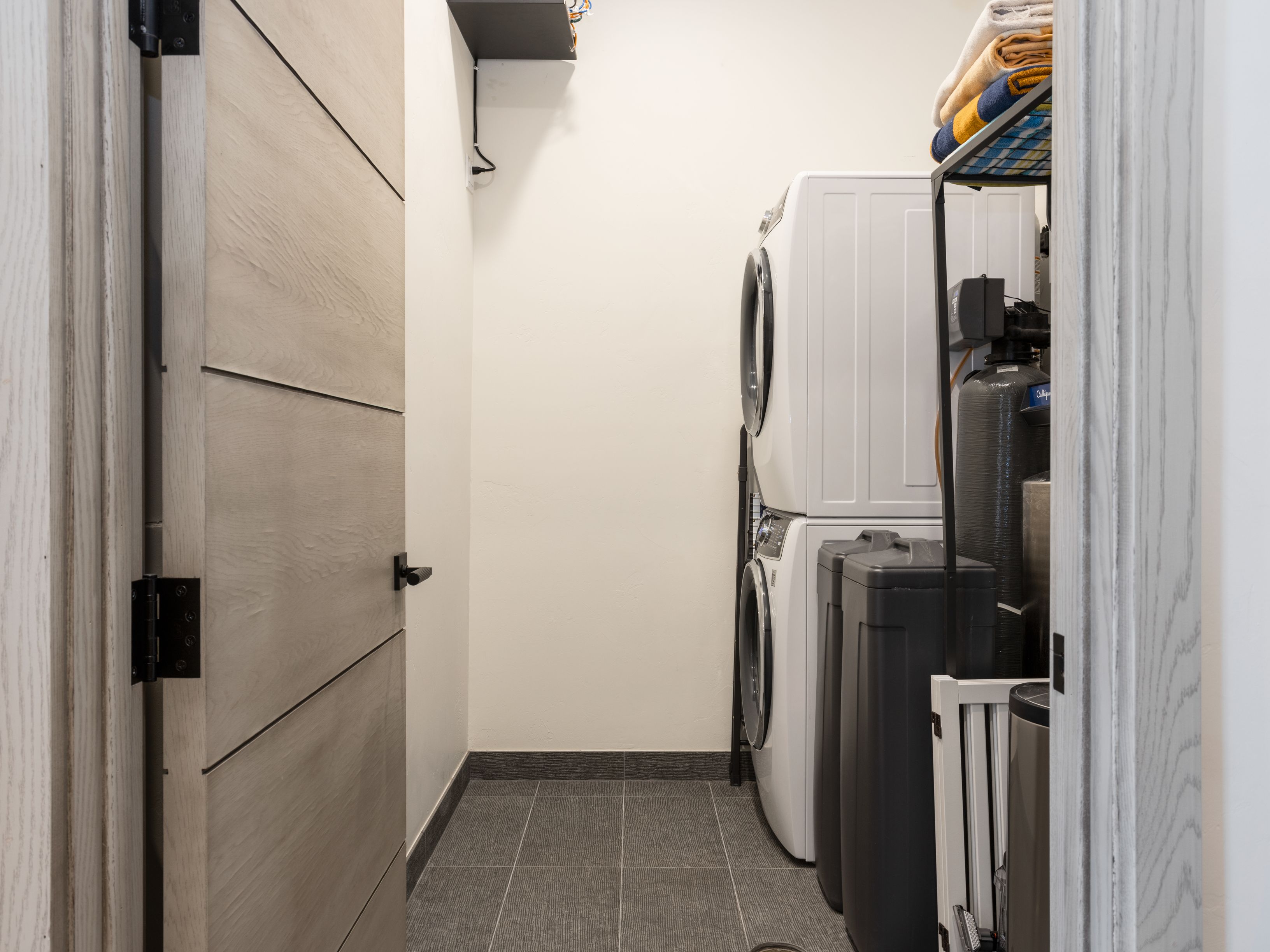 This is a well-organized laundry room featuring a stacked washer and dryer set, a water filtration system, and a storage rack with neatly folded towels. The room has a modern aesthetic with gray tiled flooring and a light-colored wooden door with black hardware. The perspective is from the doorway, offering a glimpse into the functional space.