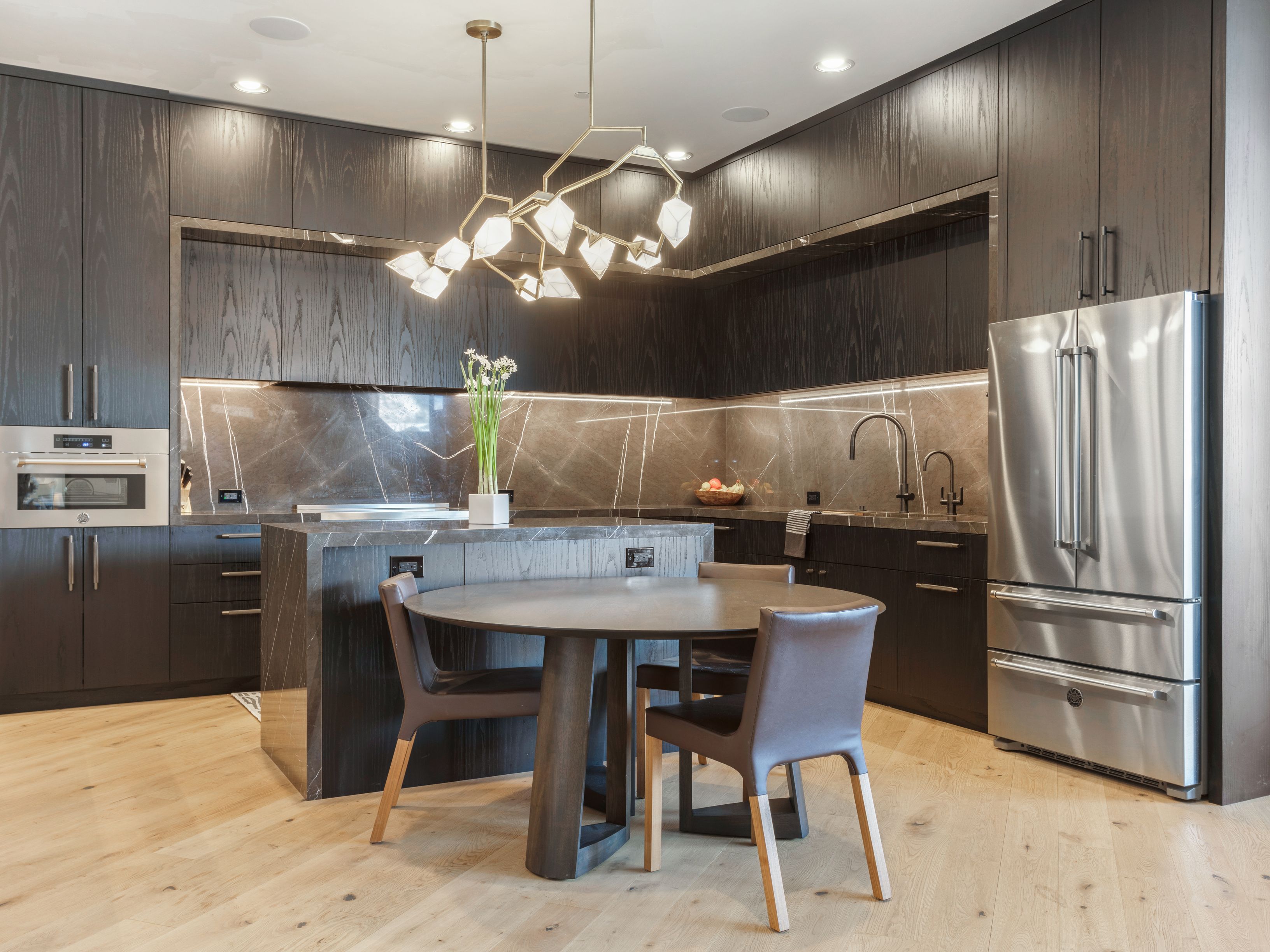 This is a modern kitchen with dark wood cabinetry and marble countertops. A unique chandelier hangs above a round dining table with chairs. Stainless steel appliances and recessed lighting add to the contemporary feel of the space.