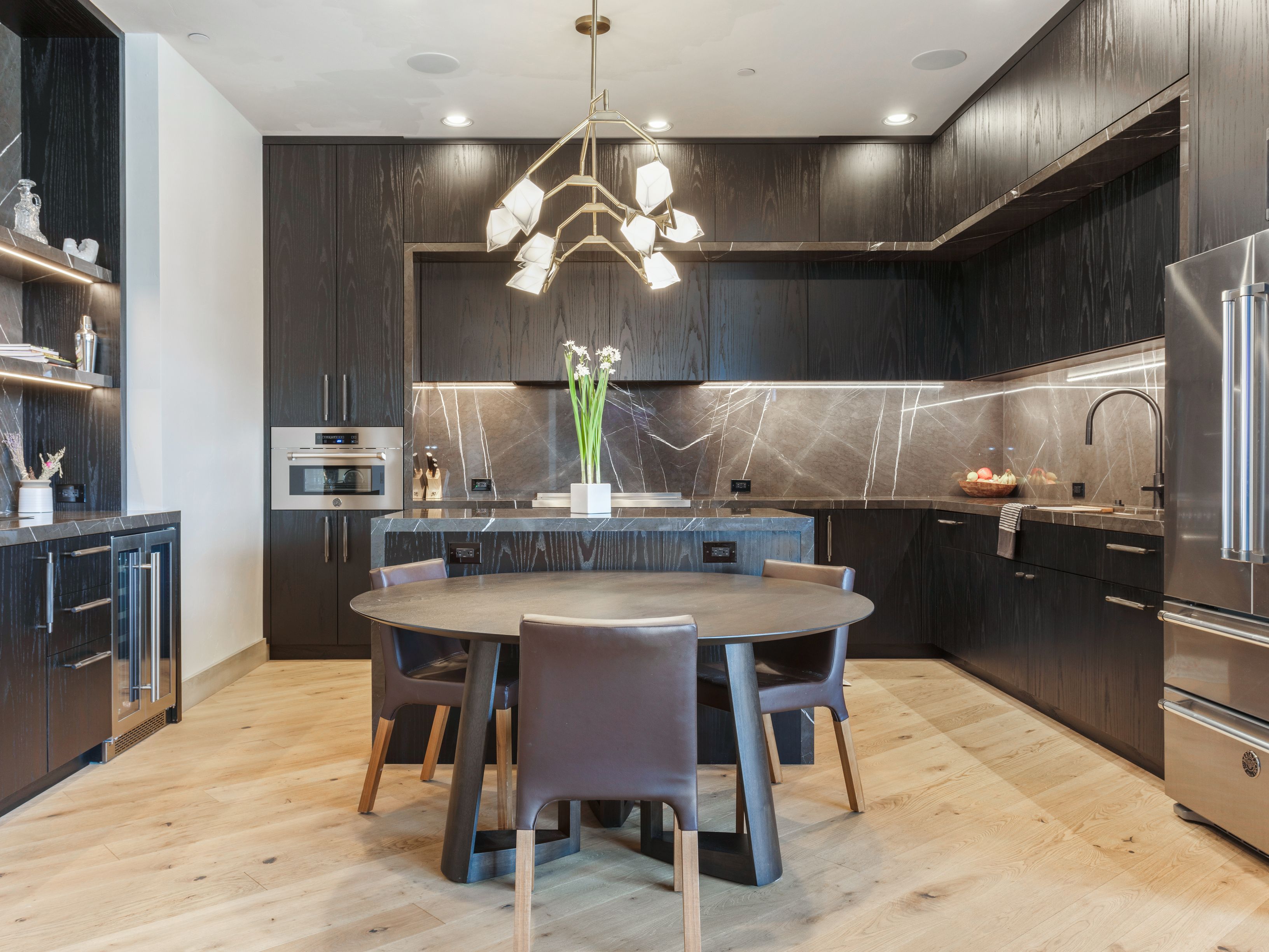 This is a modern kitchen with dark wood cabinetry and marble countertops. A round dining table with four chairs sits in the center of the room, illuminated by a unique chandelier. Stainless steel appliances and a built-in wine fridge add to the luxurious feel of the space.