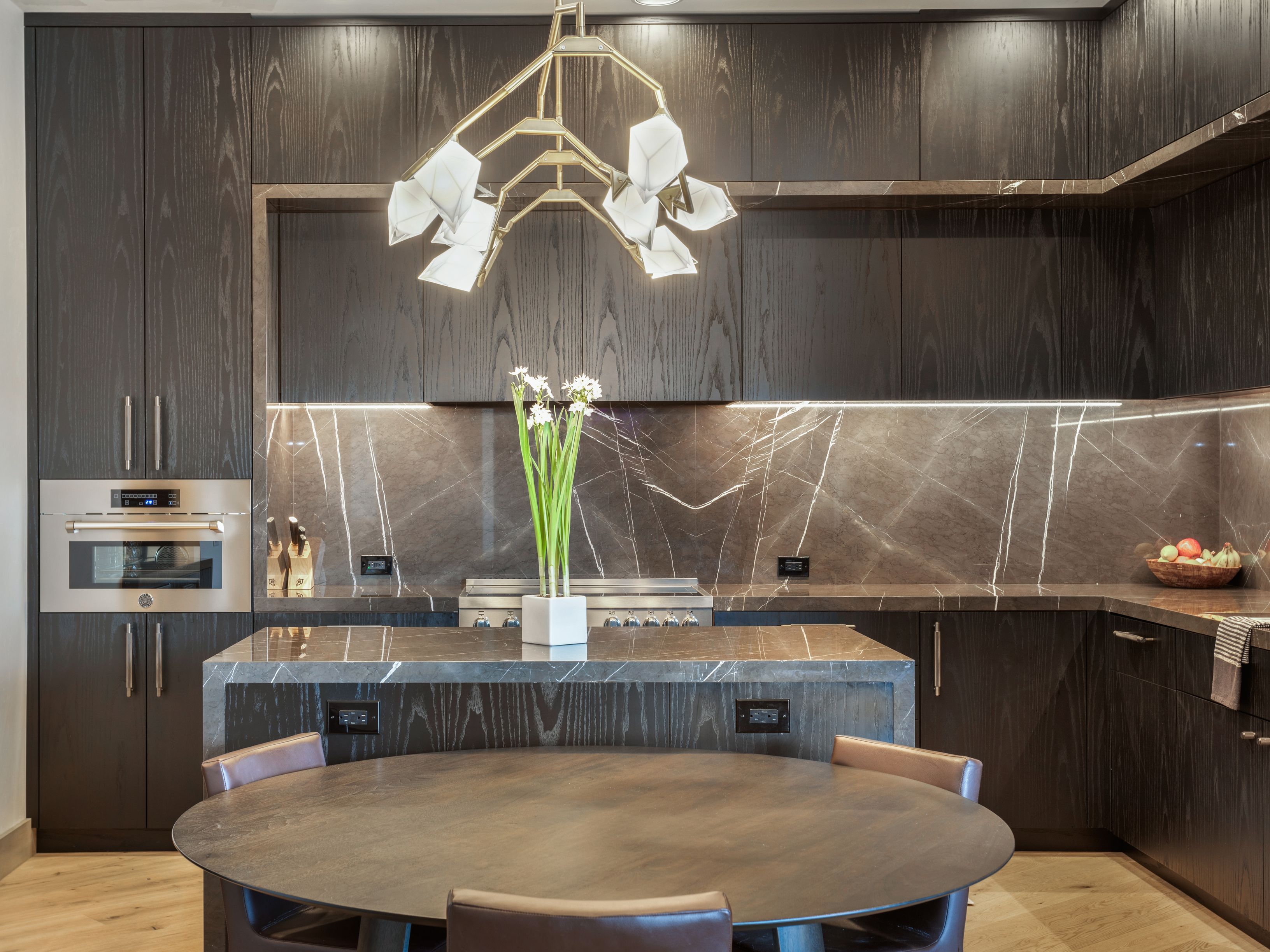 This is a modern kitchen with dark wood cabinetry and marble countertops with striking white veins. A unique chandelier hangs above a round dining table with leather chairs. Stainless steel appliances are integrated into the cabinetry, and the overall design is sleek and sophisticated.