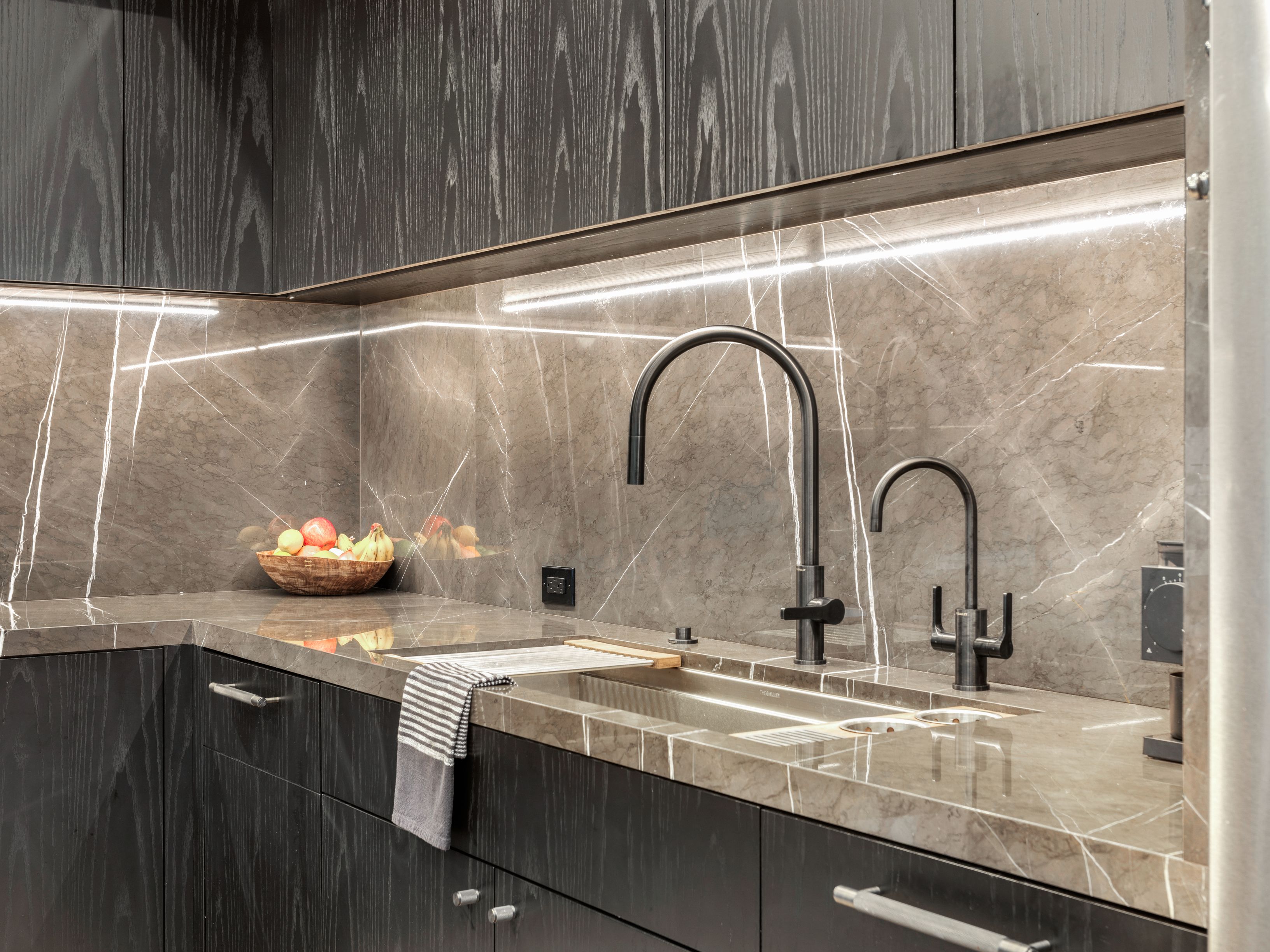 This is a modern kitchen featuring dark wood cabinetry and a marble backsplash with integrated lighting. The countertops are also marble, complementing the backsplash. Two sleek, matte black faucets are installed in the sink, adding to the contemporary aesthetic.