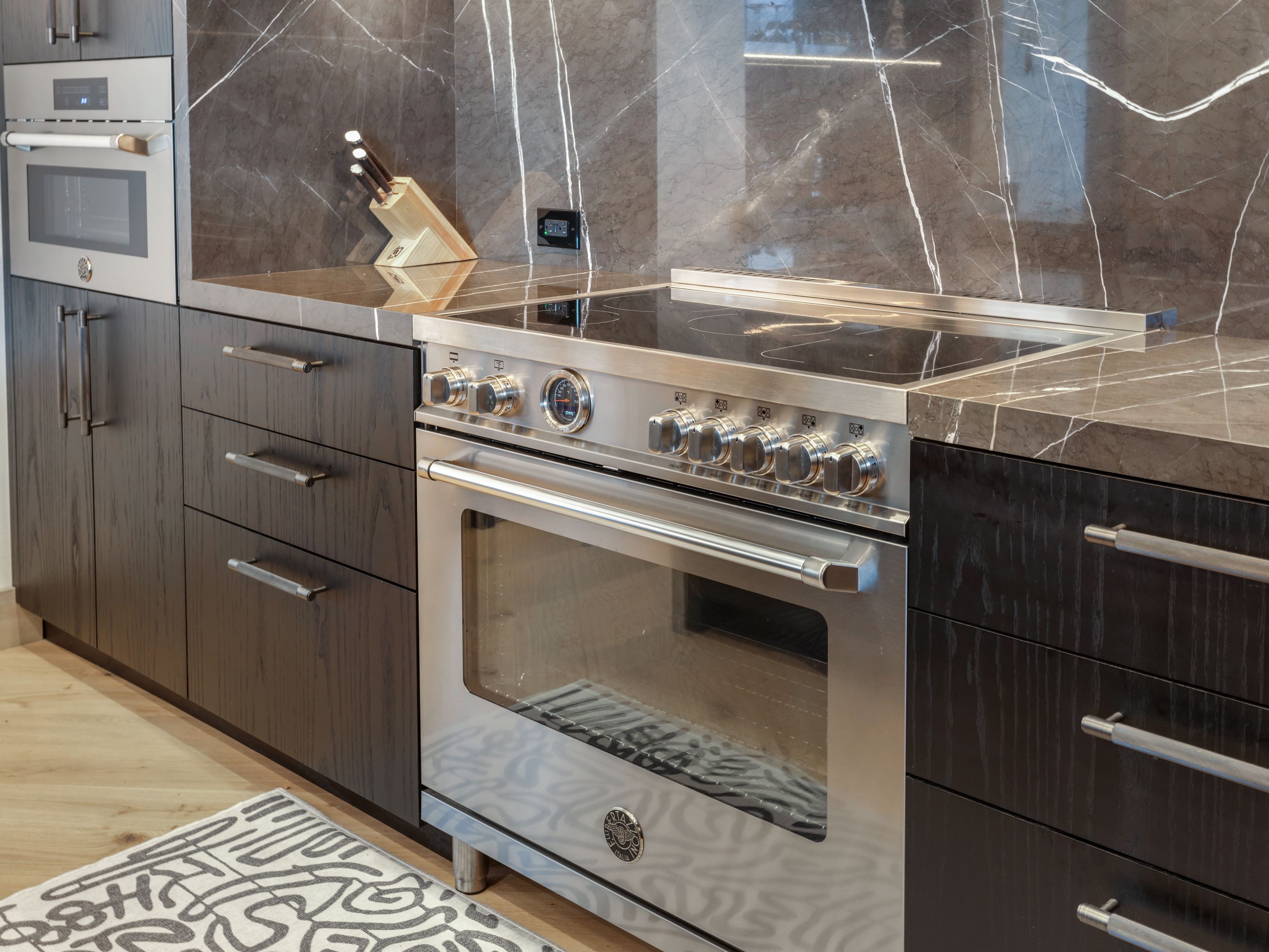This is a detailed shot of a modern kitchen featuring dark wood cabinetry with sleek, silver hardware. A stainless steel range with multiple knobs and a glass oven door is the focal point, set against a backdrop of marble backsplash and countertops. The overall impression is one of luxury and sophistication.