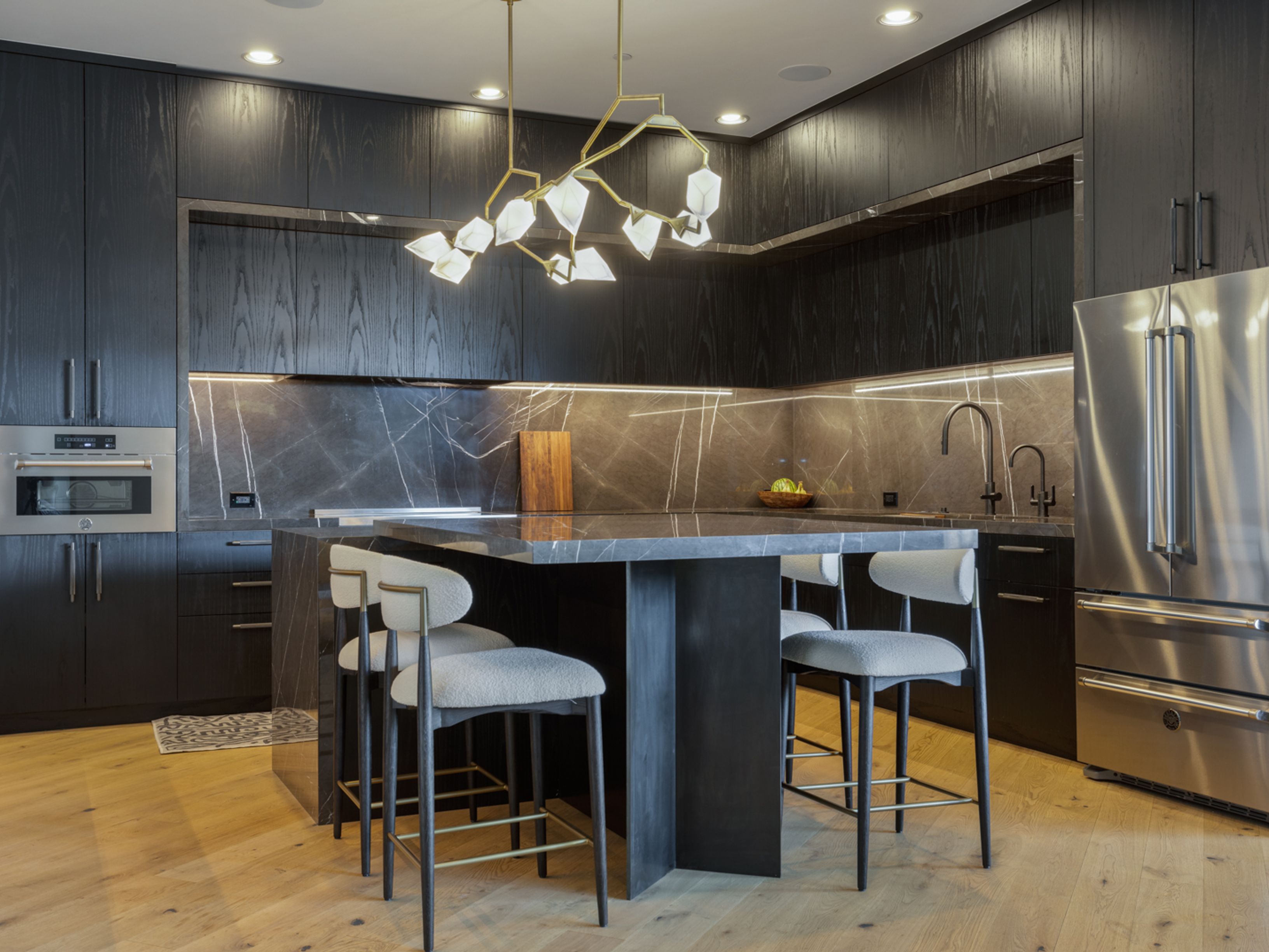 This is a modern kitchen featuring dark wood cabinetry and a marble backsplash with integrated lighting. A large island with a marble countertop and seating is centrally located, illuminated by a contemporary chandelier. Stainless steel appliances and light wood flooring complete the sleek and sophisticated design.