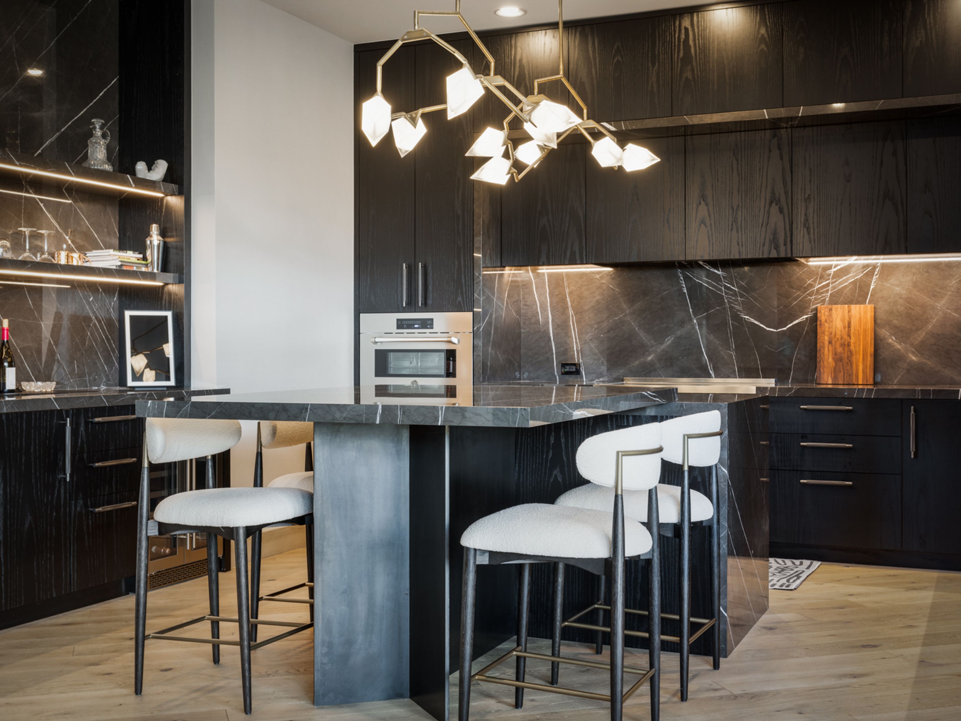 This is a modern kitchen with dark wood cabinetry and marble countertops. A large island with seating is the focal point, illuminated by a contemporary chandelier. The kitchen features high-end appliances and stylish bar stools, creating a sophisticated and inviting space.