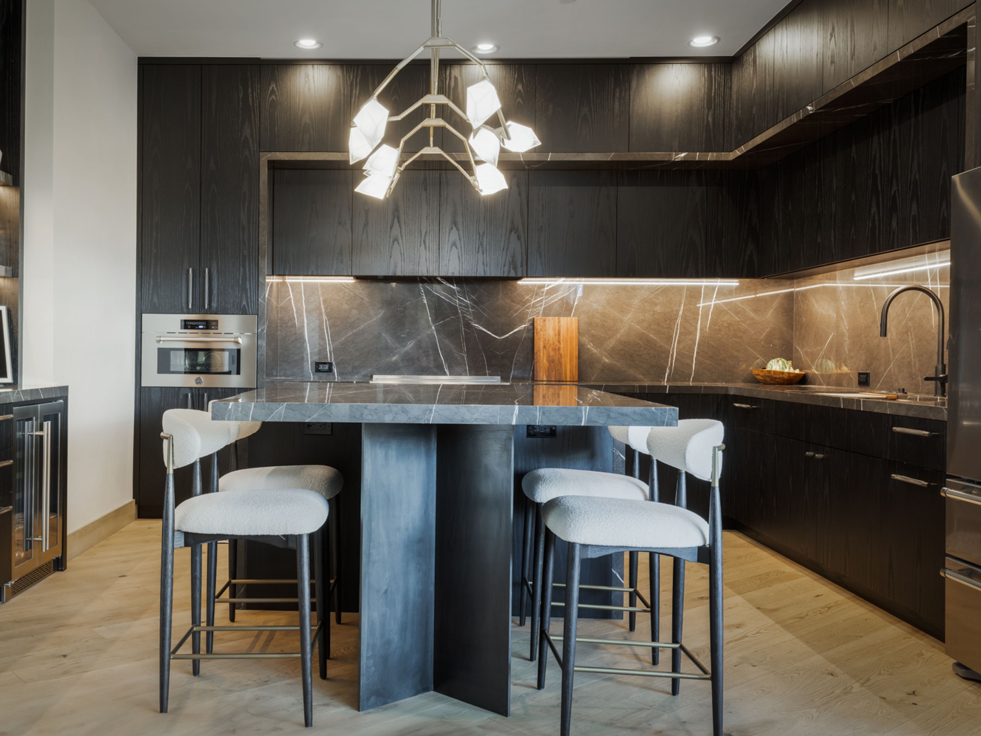 This is a modern kitchen with dark wood cabinetry and a marble backsplash with integrated lighting. A large island with a marble countertop and seating anchors the space, illuminated by a contemporary chandelier. The kitchen features stainless steel appliances and a clean, minimalist design.