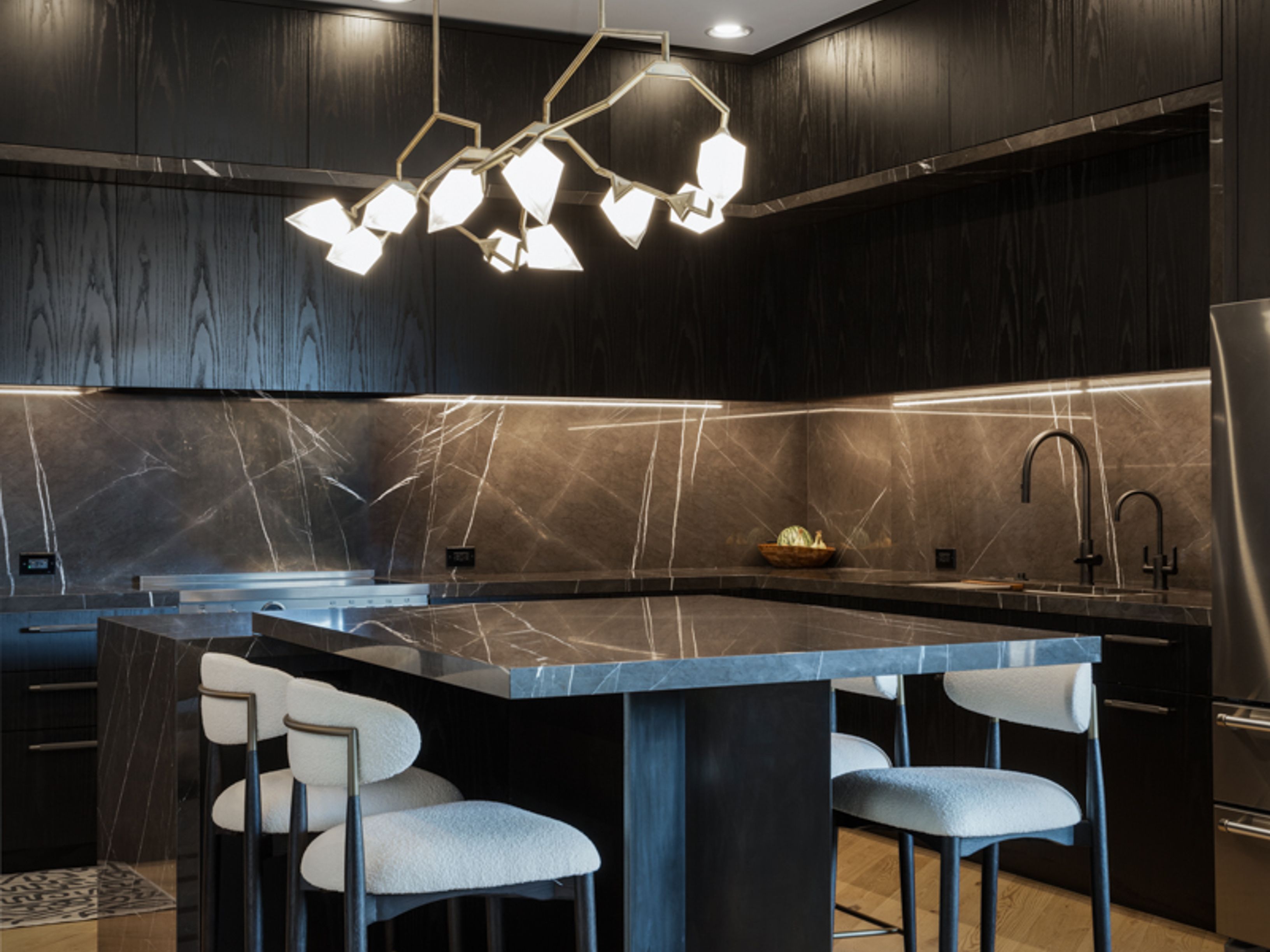 This is a modern kitchen with dark wood cabinetry and marble countertops. A unique, multi-bulb chandelier hangs above a central island with seating. The kitchen features sleek, built-in appliances and under-cabinet lighting, creating a sophisticated and luxurious atmosphere.