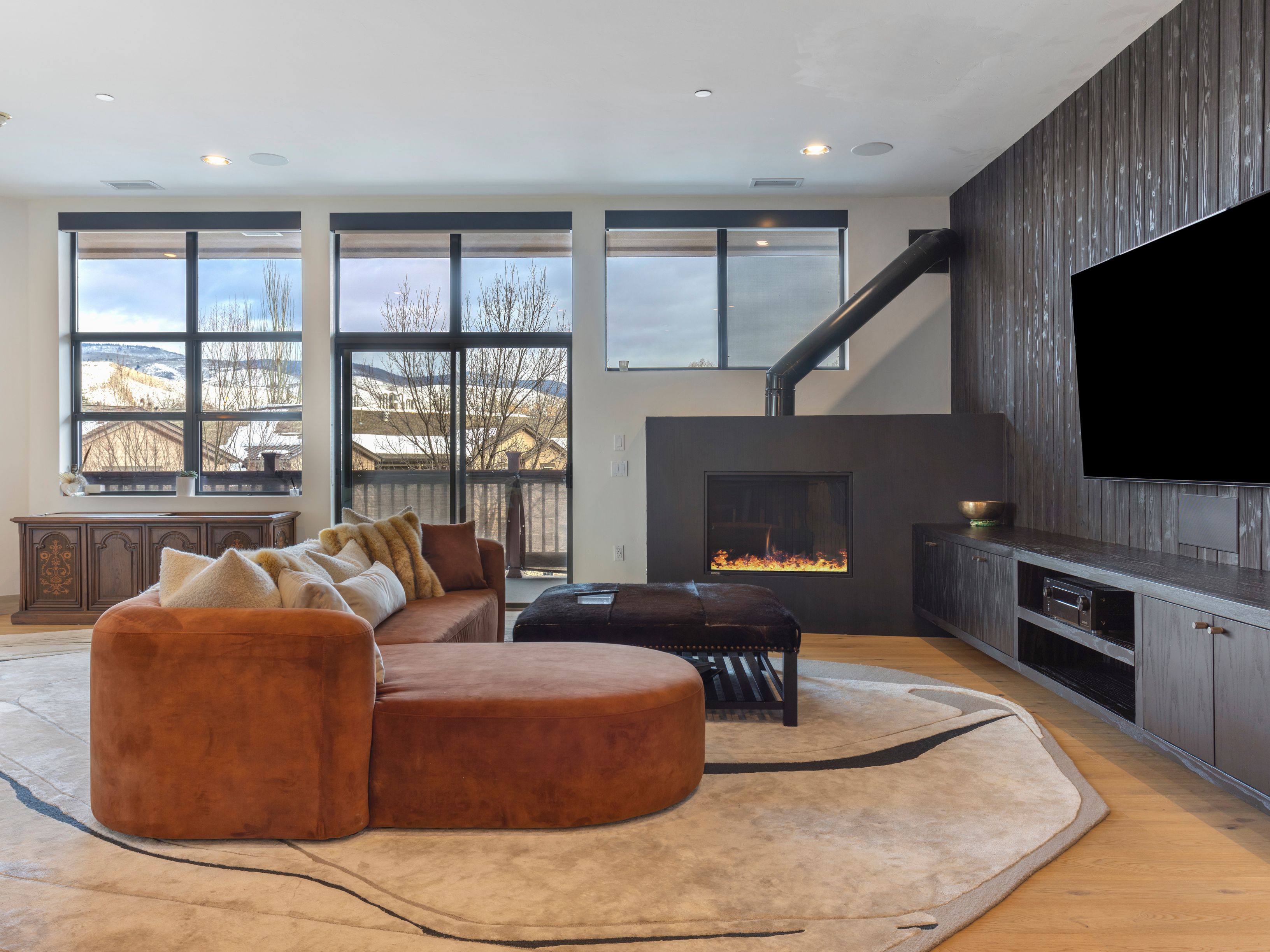 This is a modern living room featuring a brown sectional sofa with decorative pillows, a dark ottoman, and a fireplace with a black pipe extending upwards. Large windows offer a view of a snowy landscape, and a built-in entertainment center houses a large television. The room is decorated with a neutral-toned rug with a black design and has hardwood floors.