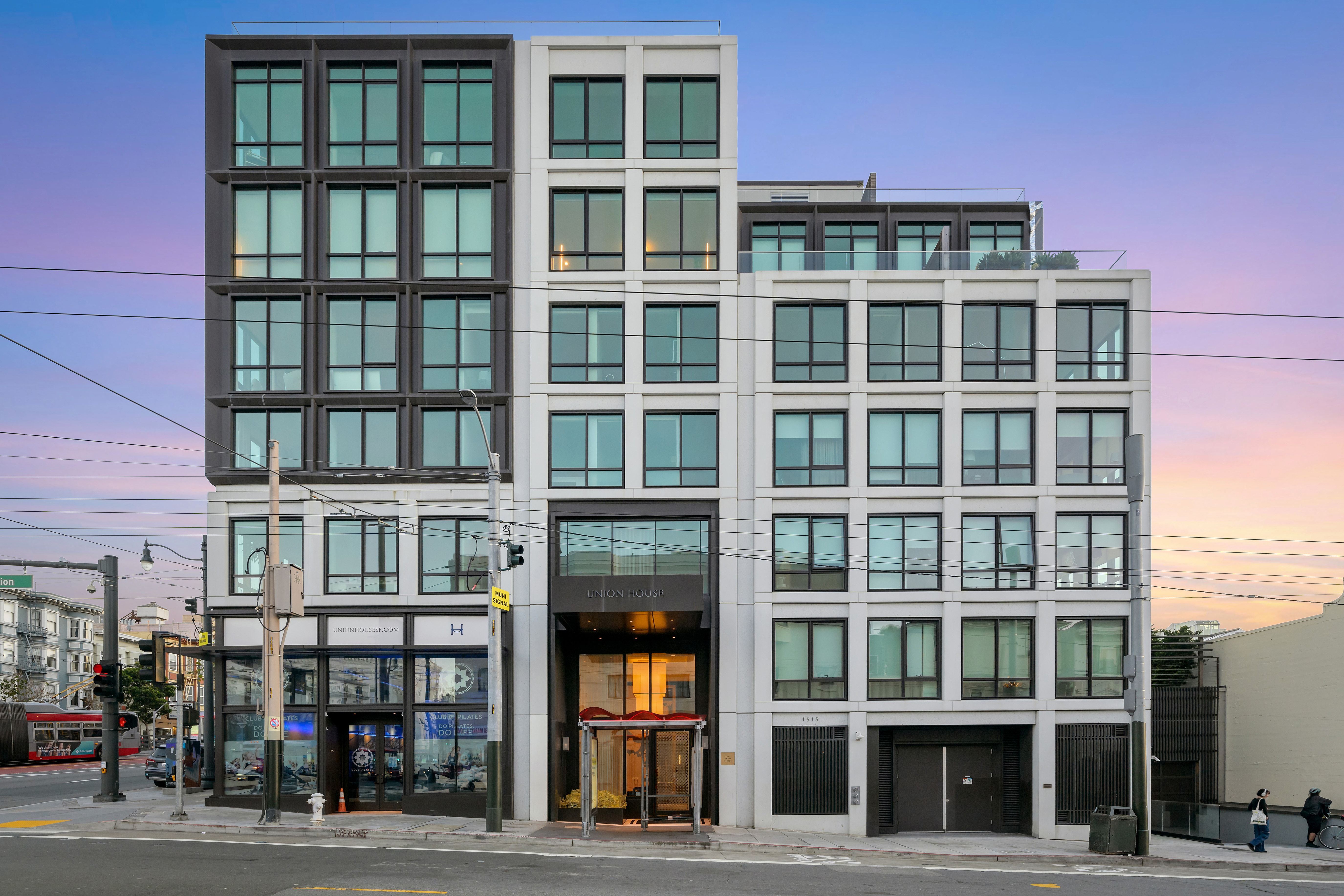 This is a striking, modern mid-rise building featuring a contemporary facade with a mix of dark-framed windows and light stone-colored cladding. The symmetrical design centers around a recessed, glass-enclosed grand entryway labeled 'Union House' and includes ground-level retail space. Its urban architectural style, clean lines, and prominent street-level presence offer a sophisticated impression ideal for premium city living.
