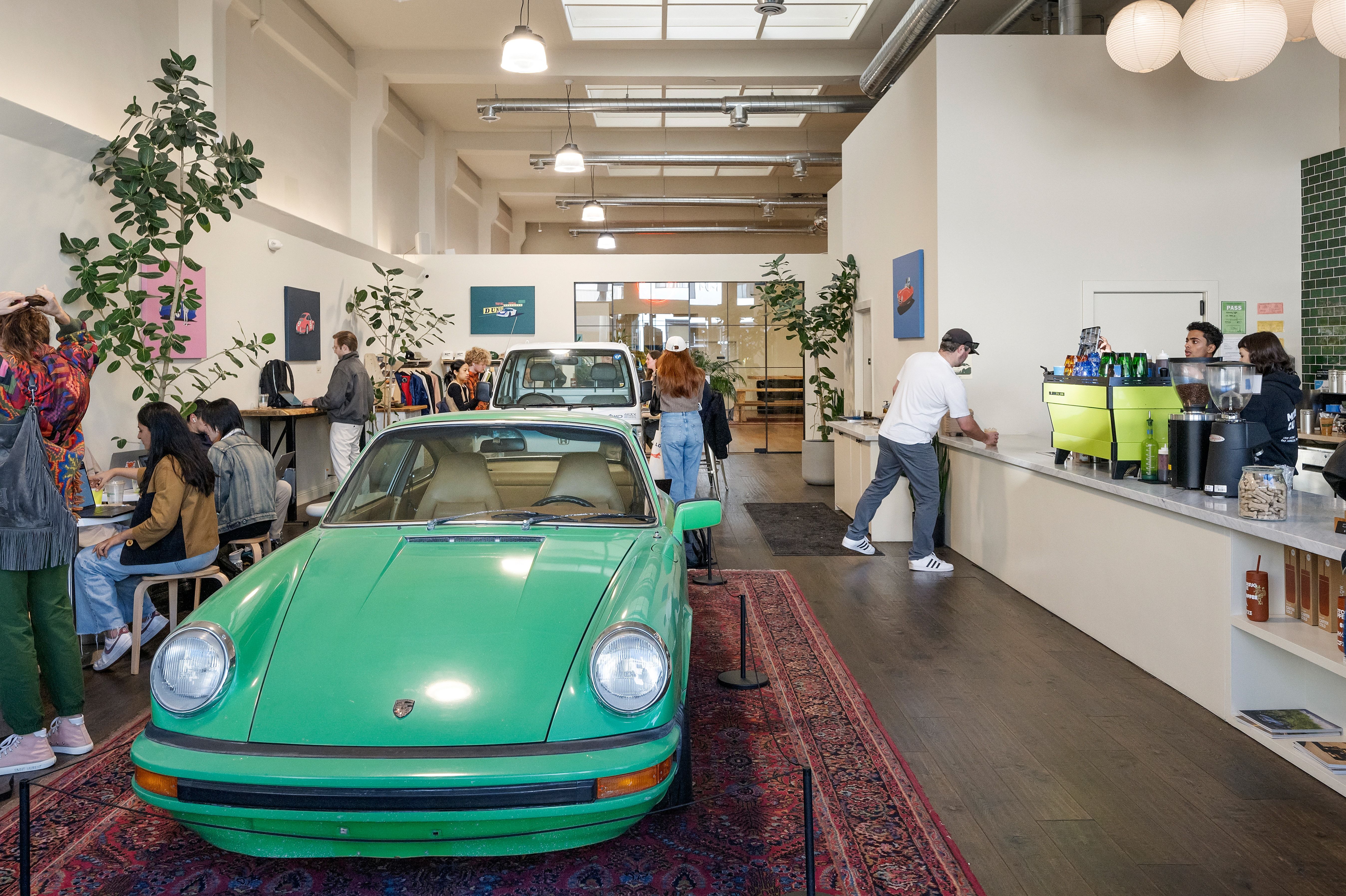 This unique, industrial-style cafe space features an open-concept layout centered around a vintage green Porsche parked prominently on a decorative area rug. The interior is characterized by high ceilings with exposed ductwork, modern pendant lighting, and a prominent coffee bar with professional espresso machinery, creating a trendy and energetic atmosphere for customers.