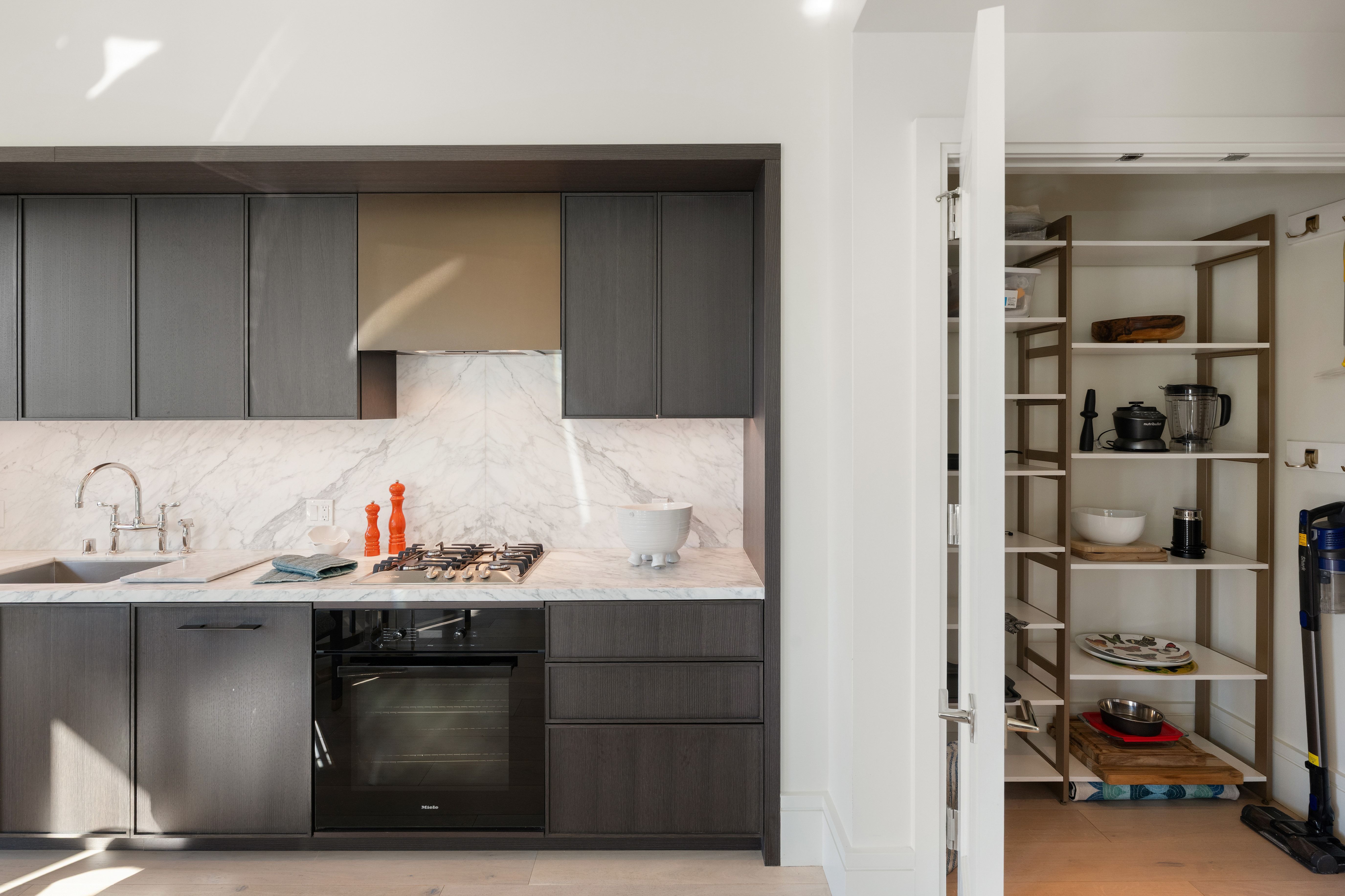 This sleek, modern kitchen features dark wood cabinetry, a striking marble backsplash, and white marble countertops, creating a sophisticated and high-end aesthetic. The space is equipped with premium integrated appliances, including a Miele oven, and flows seamlessly into a well-organized walk-in pantry on the right. Natural light highlights the premium finishes, contributing to a clean and functional design perfect for luxury living.