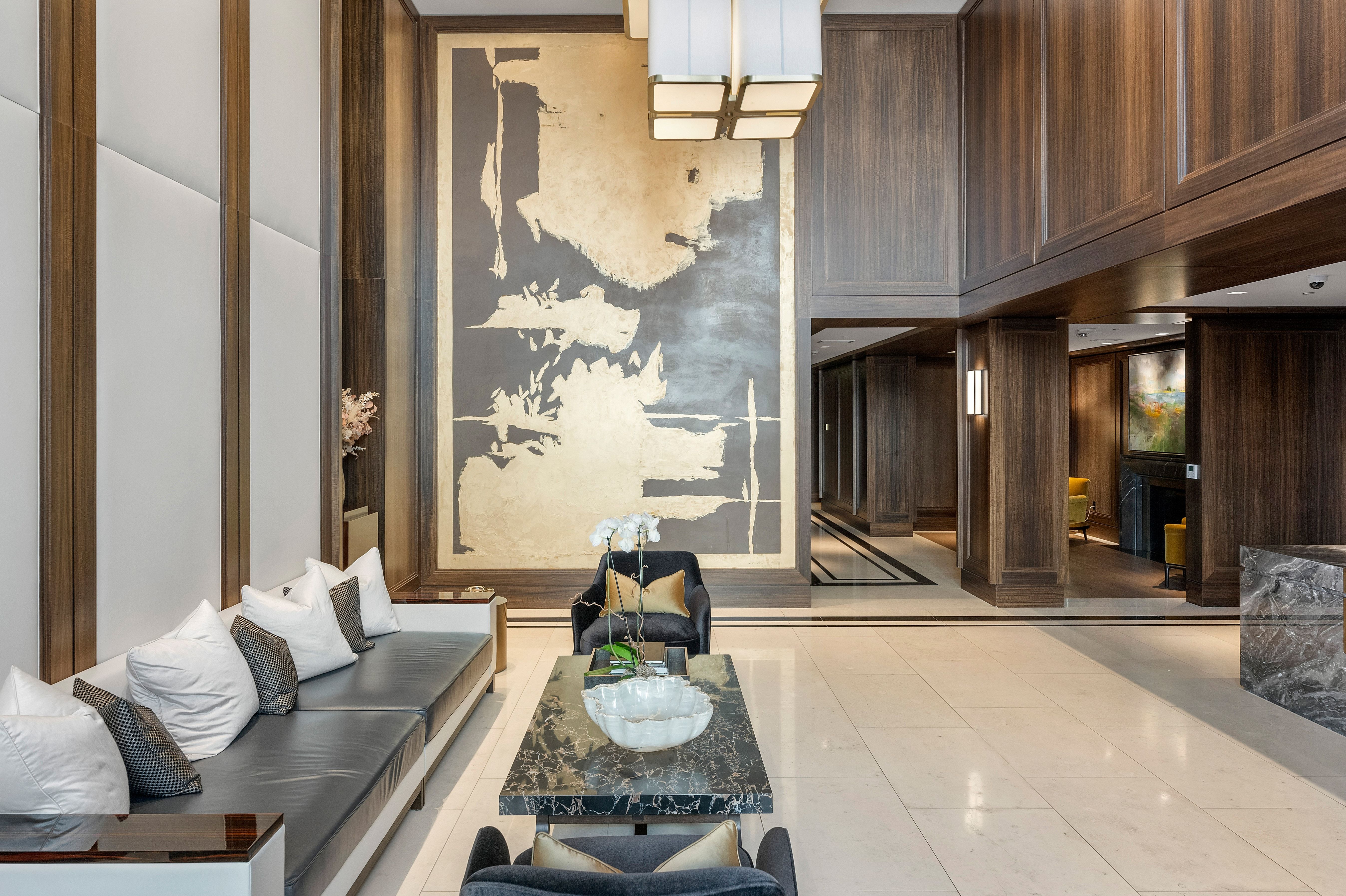 This sophisticated, high-ceilinged lobby area exudes luxury with its dark wood paneling, large-scale abstract artwork, and upscale furnishings. The space is anchored by a marble-topped coffee table and features premium finishes, creating a refined and welcoming atmosphere typical of a high-end condominium or residential complex.