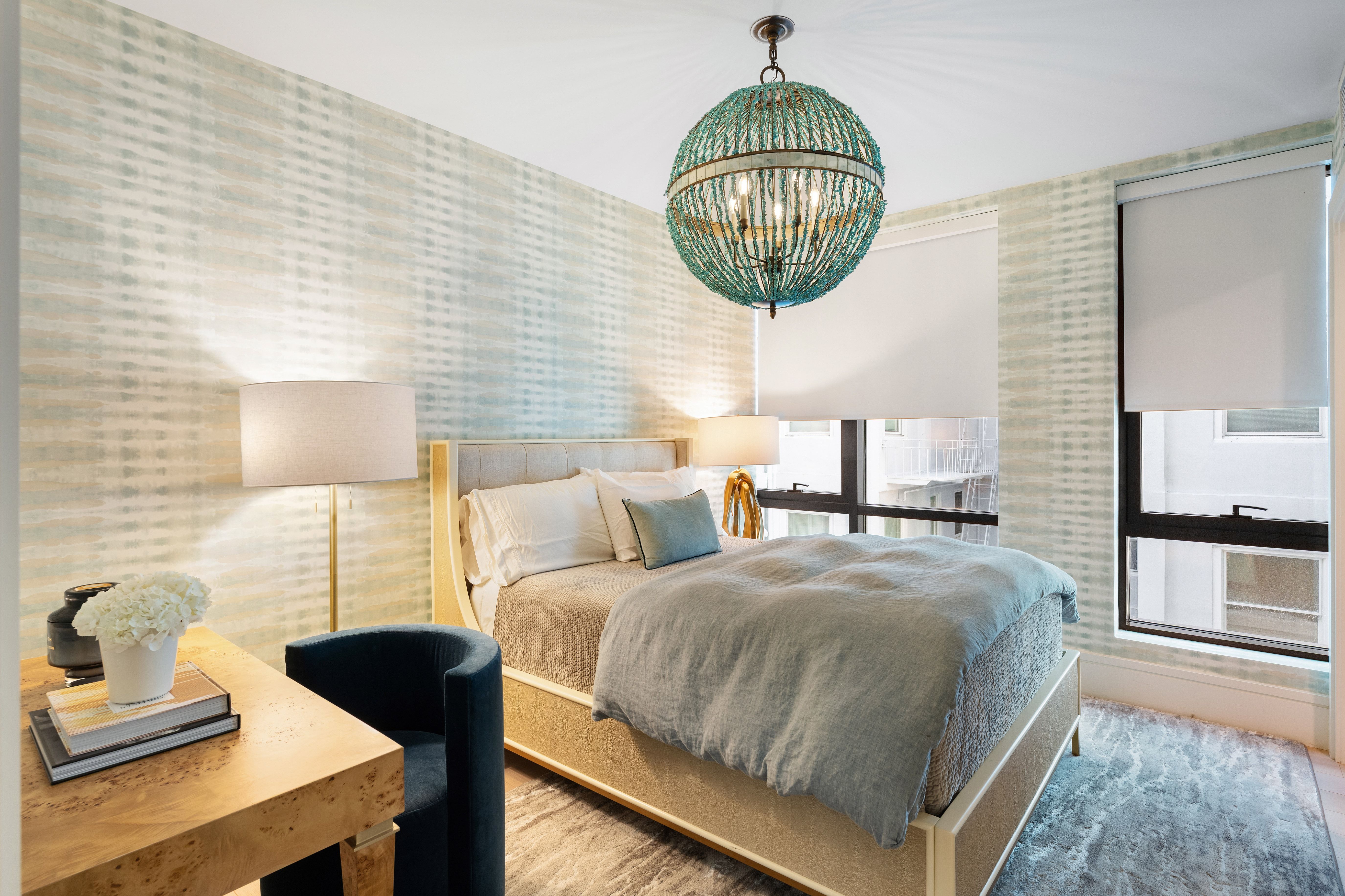 This serene bedroom features a contemporary design highlighted by a striking turquoise beaded orb chandelier and textured, soft-blue patterned wallpaper. The space includes a comfortable platform bed with light-toned bedding, a wooden desk area, and a plush navy accent chair, creating a cozy and stylish retreat. Large windows with neutral roller shades allow for natural light, making the room feel bright and inviting.