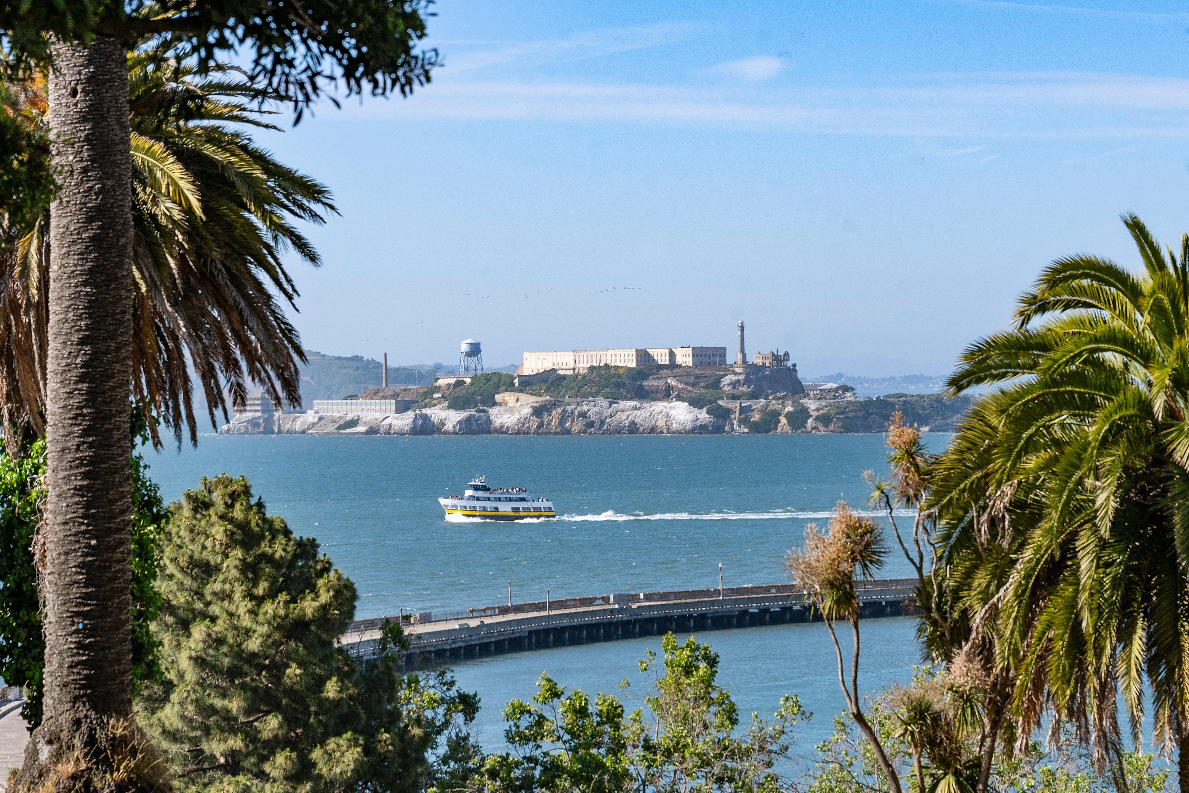 This image features a scenic view of Alcatraz Island from a vantage point framed by palm trees and foliage. A ferry boat travels across the water in front of the island, which is connected to a pier extending into the bay. The composition emphasizes a desirable coastal location and provides a picturesque perspective often highlighted in listings for waterfront properties.