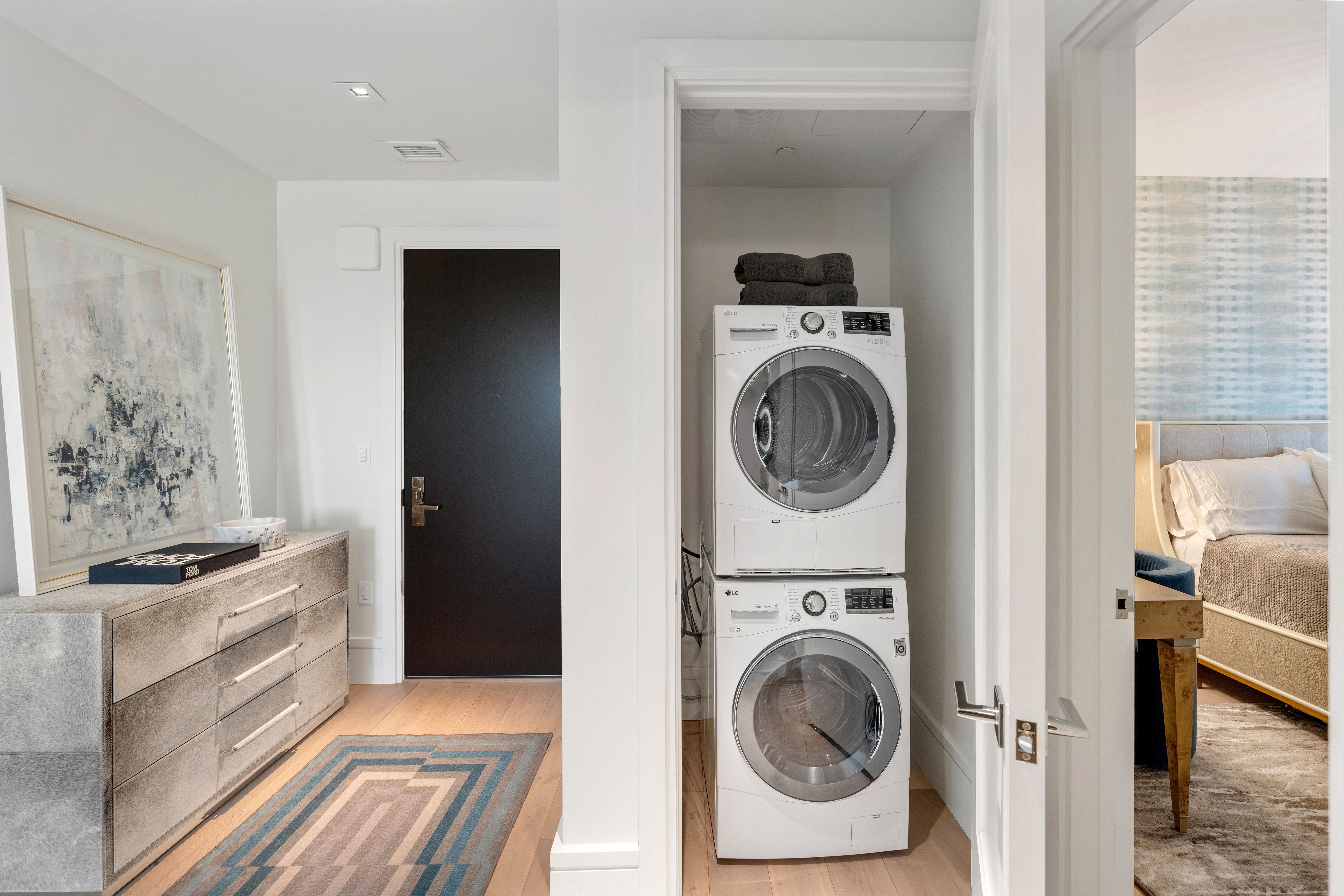This interior view showcases a discreet, stacked LG washer and dryer unit housed within a functional laundry closet. The adjacent hallway features a stylish textured dresser, modern artwork, and a geometric rug, leading towards a bedroom visible through an open doorway. The space exhibits a clean, contemporary design with neutral tones and efficient use of space.