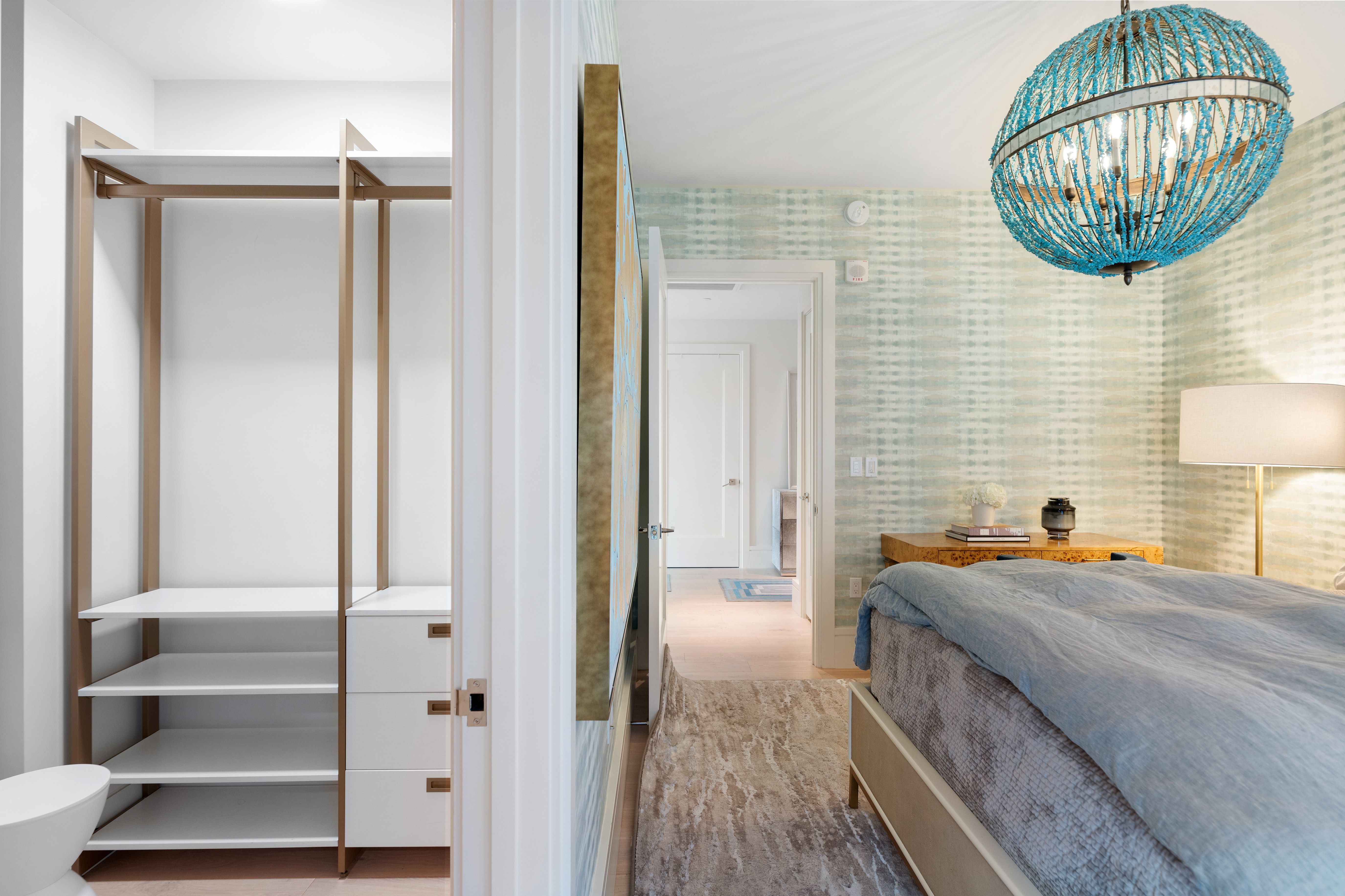 This elegant bedroom features a striking blue beaded chandelier, light green patterned wallpaper, and a cozy bed with layered textiles. The room flows seamlessly into an organized open walk-in closet with white shelving and gold-toned finishes, showcasing both functional storage and sophisticated design.