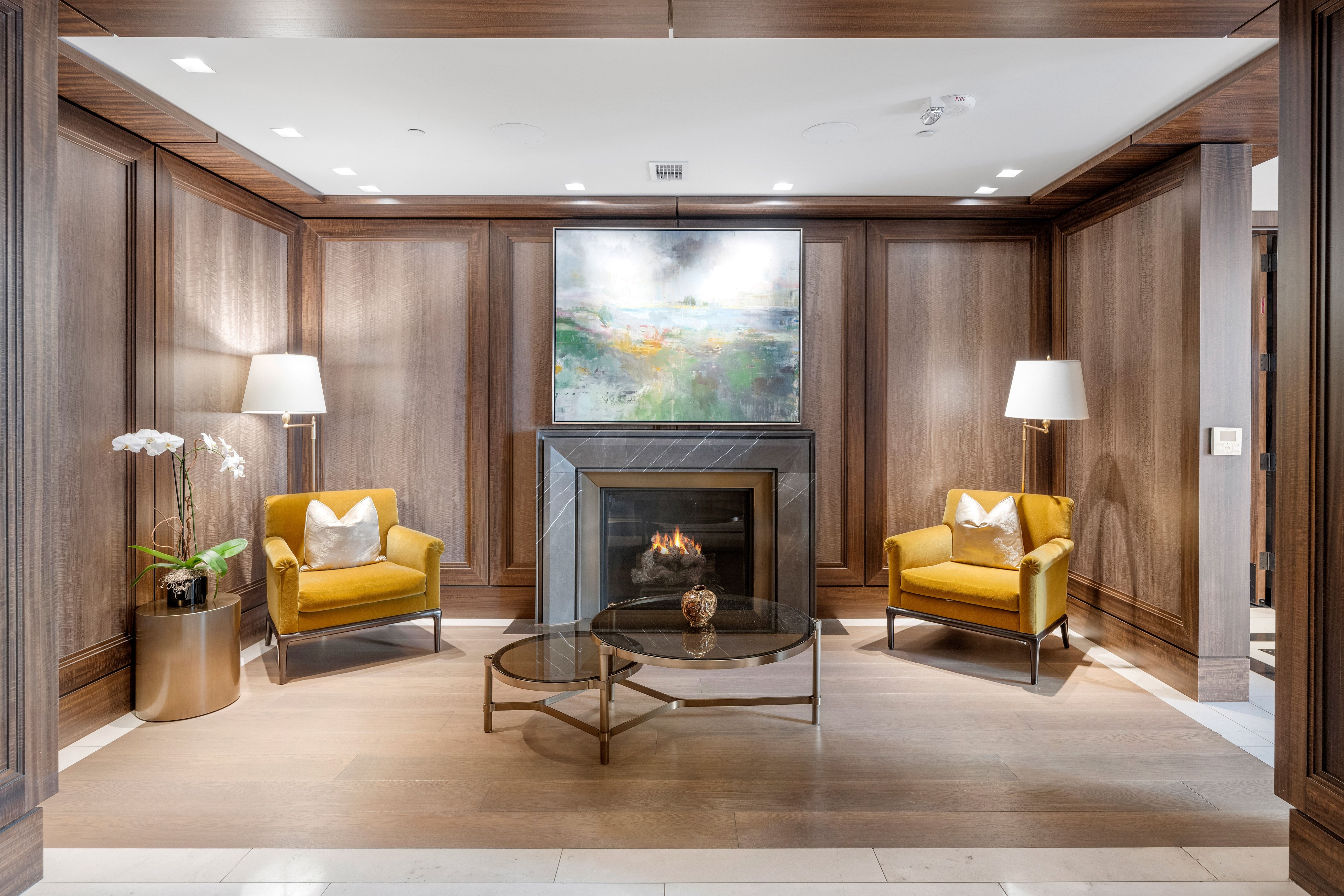 This refined lounge area, likely part of a residential clubhouse or building common space, features exquisite wood-paneled walls and a sophisticated stone fireplace as its focal point. The room is furnished with two elegant mustard-colored armchairs, a glass-topped nesting coffee table, and a pedestal planter with white orchids, creating a welcoming and upscale atmosphere. The space blend of classic millwork and modern furniture provides a luxurious and comfortable environment for residents.
