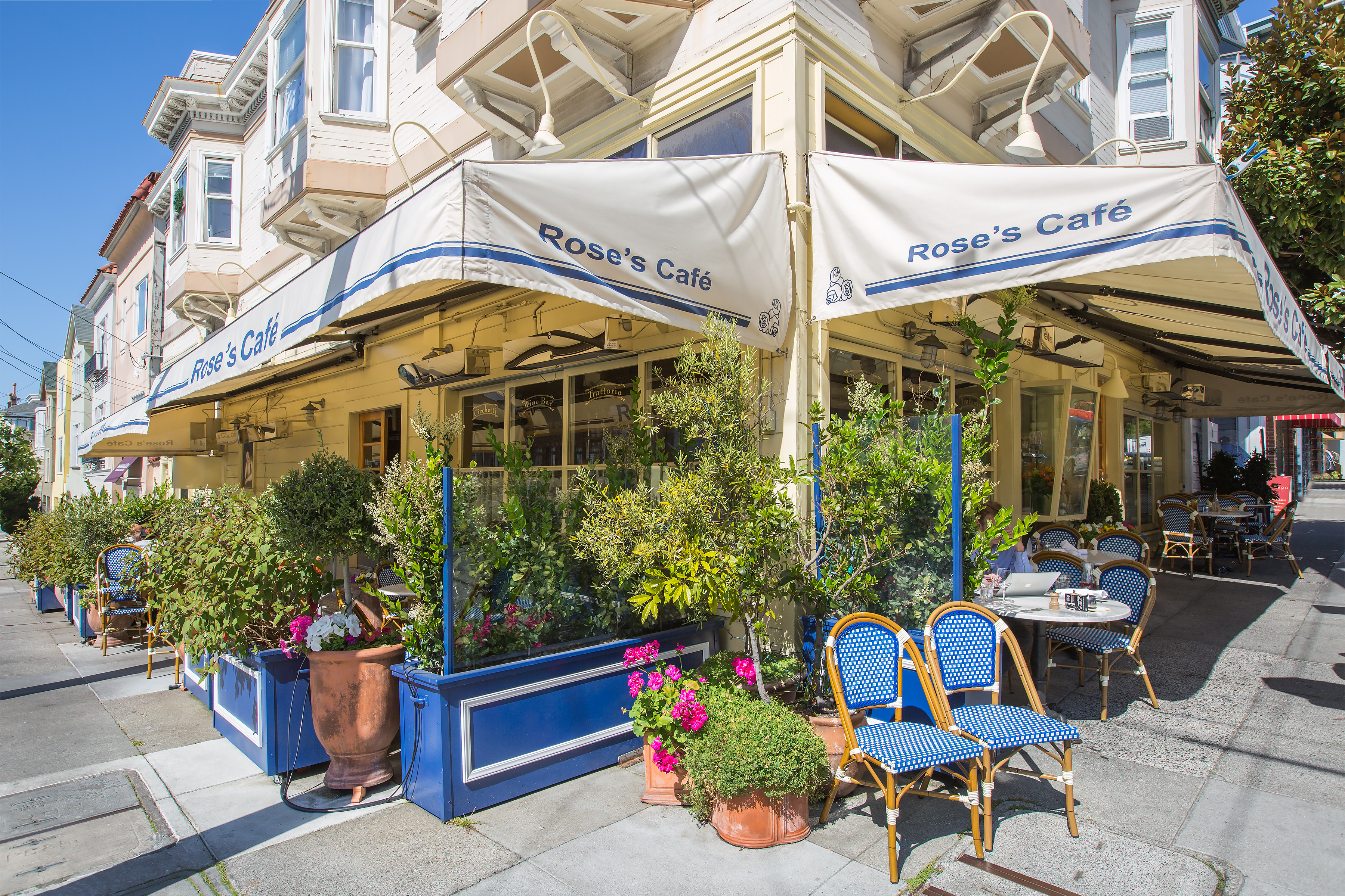 This image showcases the inviting corner storefront of 'Rose's Café,' featuring a classic San Francisco neighborhood aesthetic with wooden siding and a wraparound outdoor dining area. The space is adorned with charming blue and white bistro-style chairs, lush potted greenery, and vibrant flowering plants that create an appealing sidewalk atmosphere. Warm lighting and crisp branding on the awnings contribute to the building's highly walkable, community-oriented appeal.