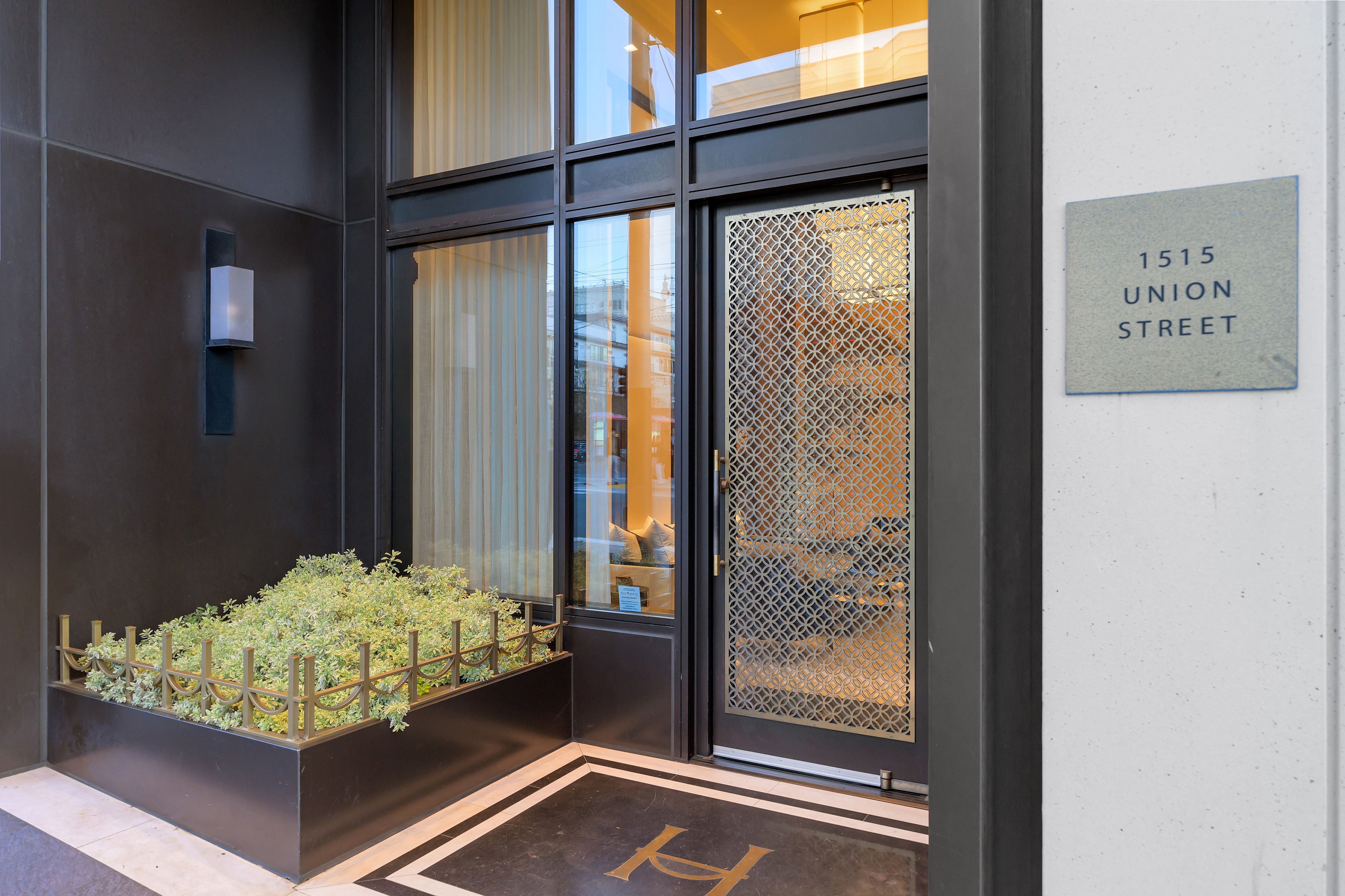 This sophisticated entryway features a decorative, gold-toned metal screen door set against a sleek, dark-framed glass facade. To the left, a modern planter box with lush greenery sits atop dark marble flooring, while a minimalist wall-mounted light fixture adds a touch of elegance. A plaque indicating the address '1515 Union Street' is prominently displayed on the adjacent white marble wall, contributing to the building's upscale and professional curb appeal.