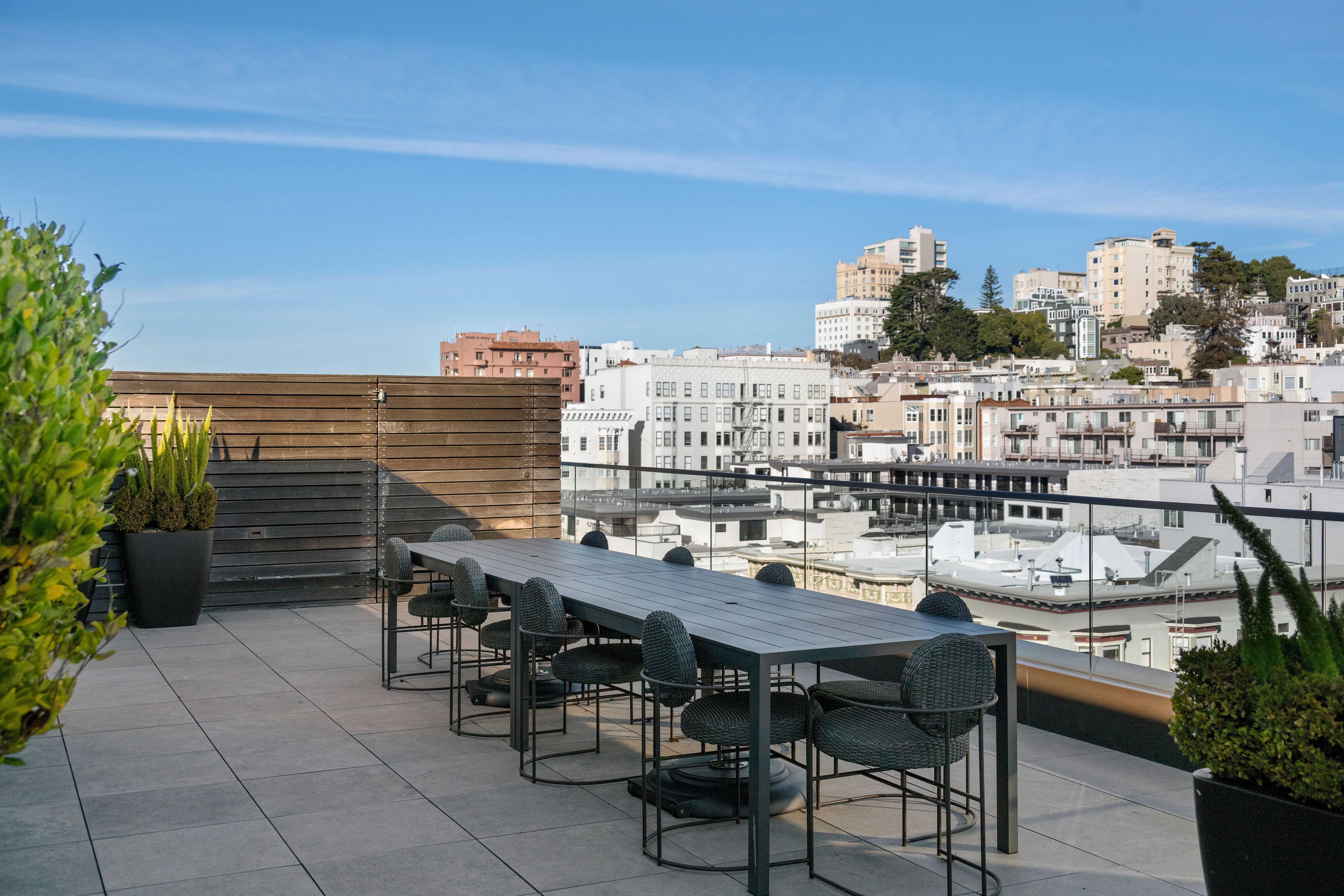 This rooftop deck features a sleek, long dark dining table surrounded by matching contemporary outdoor chairs, perfect for alfresco entertaining. The space includes a privacy-enhancing wooden screen and potted greenery, all set against a stunning urban skyline backdrop. The modern design and panoramic city views create an inviting atmosphere for relaxation or social gatherings.