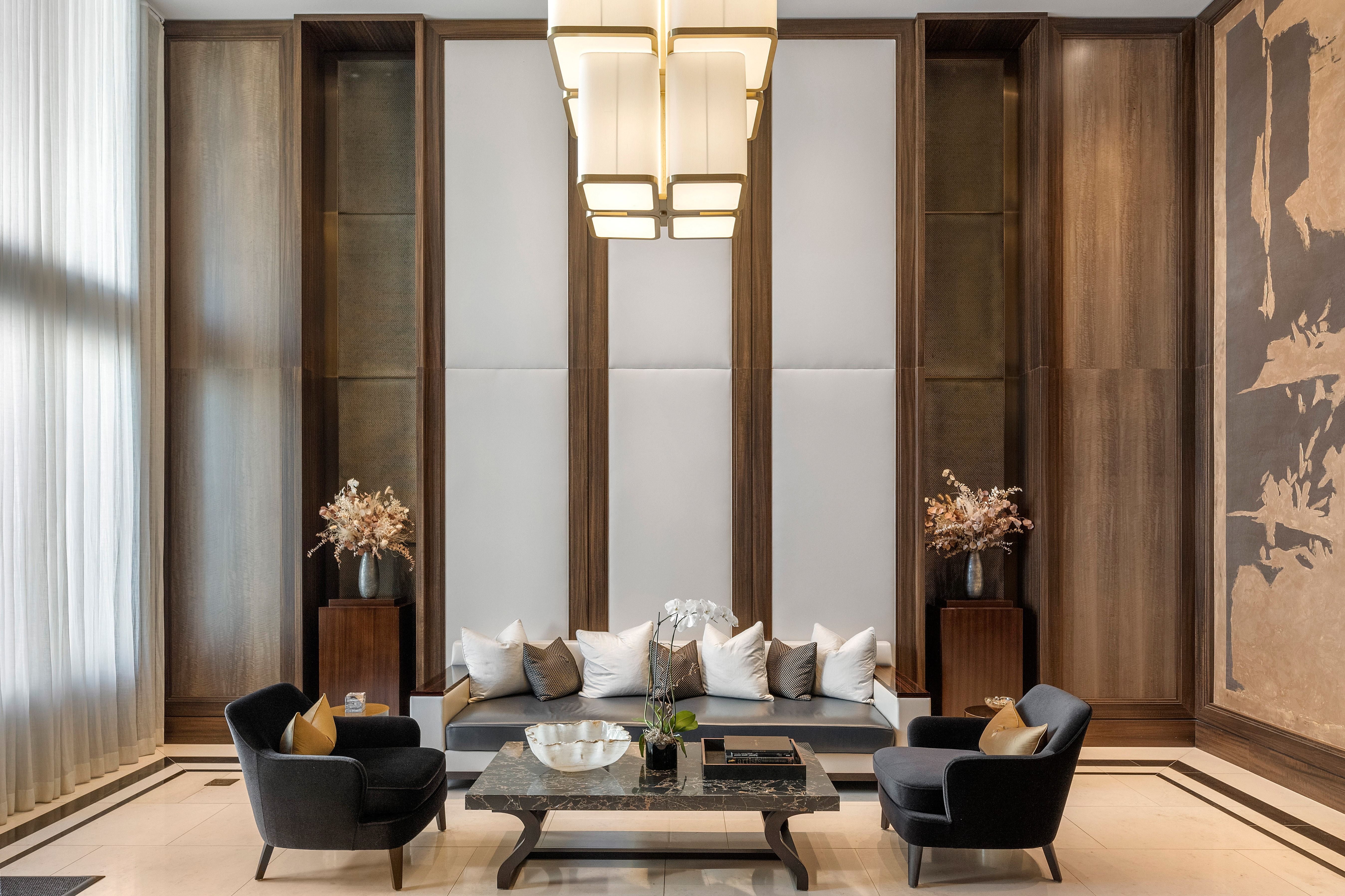 This elegant and sophisticated living room area showcases a modern, high-end design with a symmetrical layout. Key features include an expansive focal wall with wood-paneled accents, plush charcoal armchairs, a central sofa adorned with decorative pillows, and a luxurious marble-topped coffee table. The space is illuminated by a striking, contemporary chandelier and accented by floral arrangements, creating a refined and welcoming atmosphere suitable for a luxury residence or upscale building lounge.
