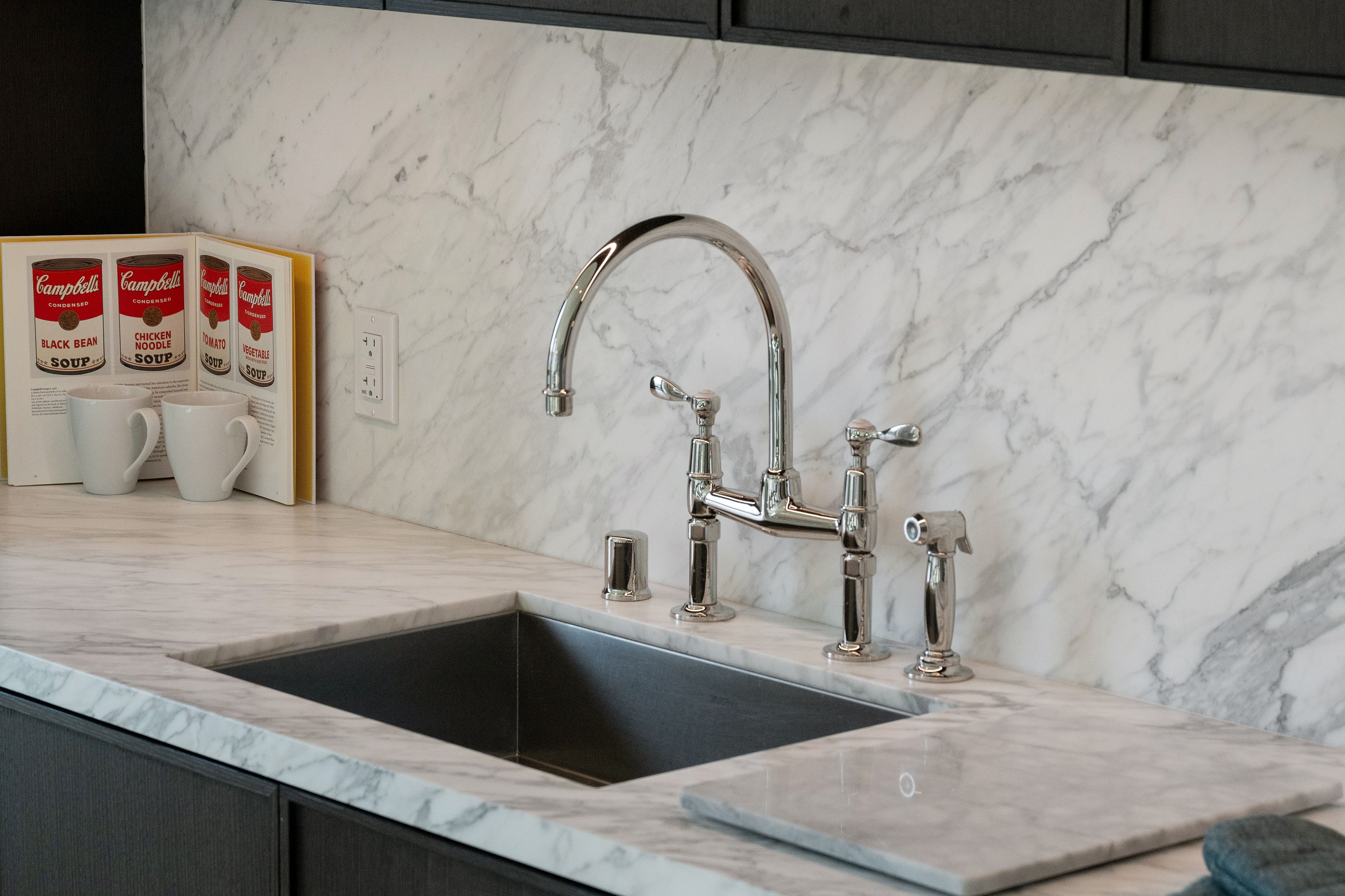 This kitchen area features high-end marble countertops and backsplash, showcasing a elegant chrome bridge-style faucet and a deep undermount sink. A cookbook featuring Campbell's soup cans adds a decorative, playful touch against the sophisticated stone surfaces. The clean, polished aesthetic highlights the premium finishes and attention to detail present in the design.