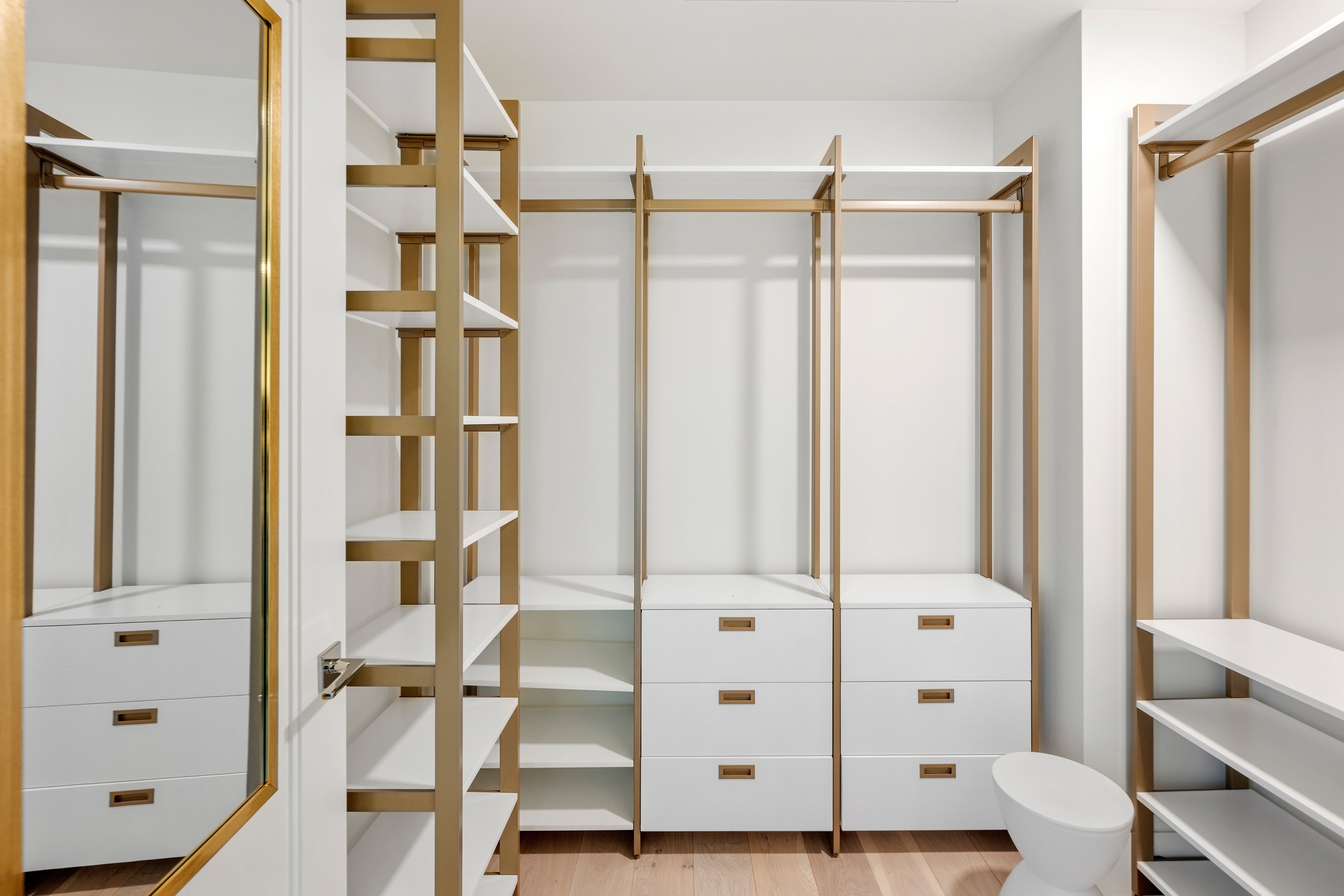 This luxurious walk-in closet features contemporary white shelving units accented with elegant gold-tone metal frames. The space includes a combination of hanging rods, open shelving, and built-in drawers with modern hardware, all set against a clean white backdrop. A large, gold-framed mirror on the left adds sophistication while creating a spacious and bright aesthetic ideal for organized storage.