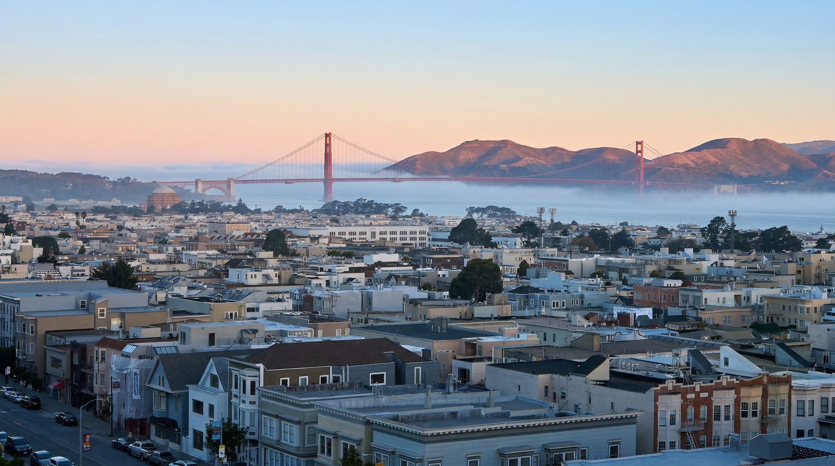 This panoramic aerial view showcases a dense urban neighborhood in San Francisco with a majestic outlook towards the Golden Gate Bridge. The scene captures the city's signature architectural landscape of multi-story buildings and historic residential housing set against a soft, sunset-lit sky and rolling coastal hills. The pervasive morning fog adds a dramatic, ethereal quality, highlighting the iconic bridge's structure and the unique appeal of this city location.