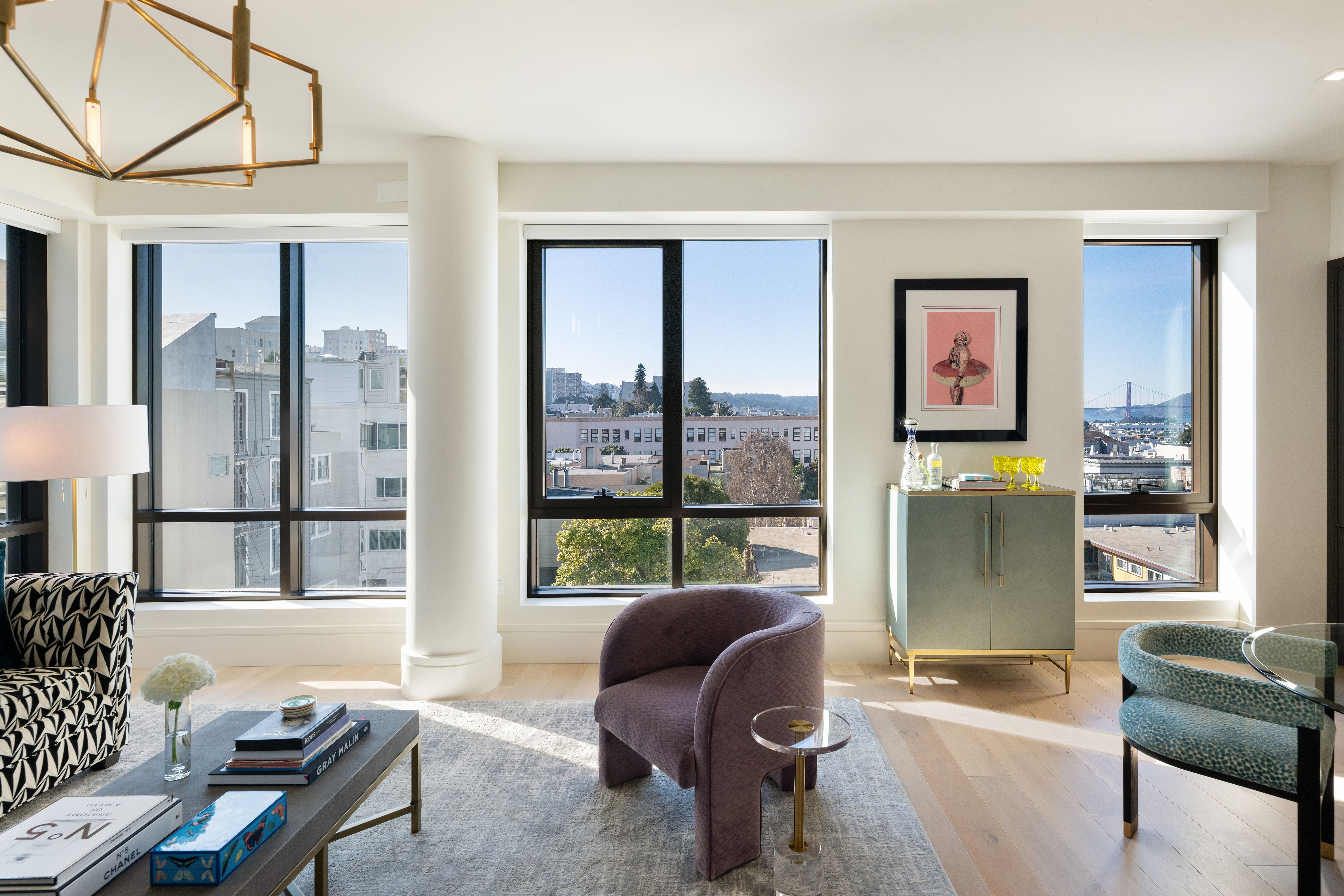 This modern, light-filled living room showcases a sophisticated blend of textures and colors, highlighted by floor-to-ceiling windows with expansive city and Golden Gate Bridge views. The space features a distinctive mauve accent chair, a patterned living room set, and a contemporary sideboard, all anchored by a soft gray area rug. Elegant lighting fixtures and curated decor details contribute to a chic and upscale urban aesthetic.