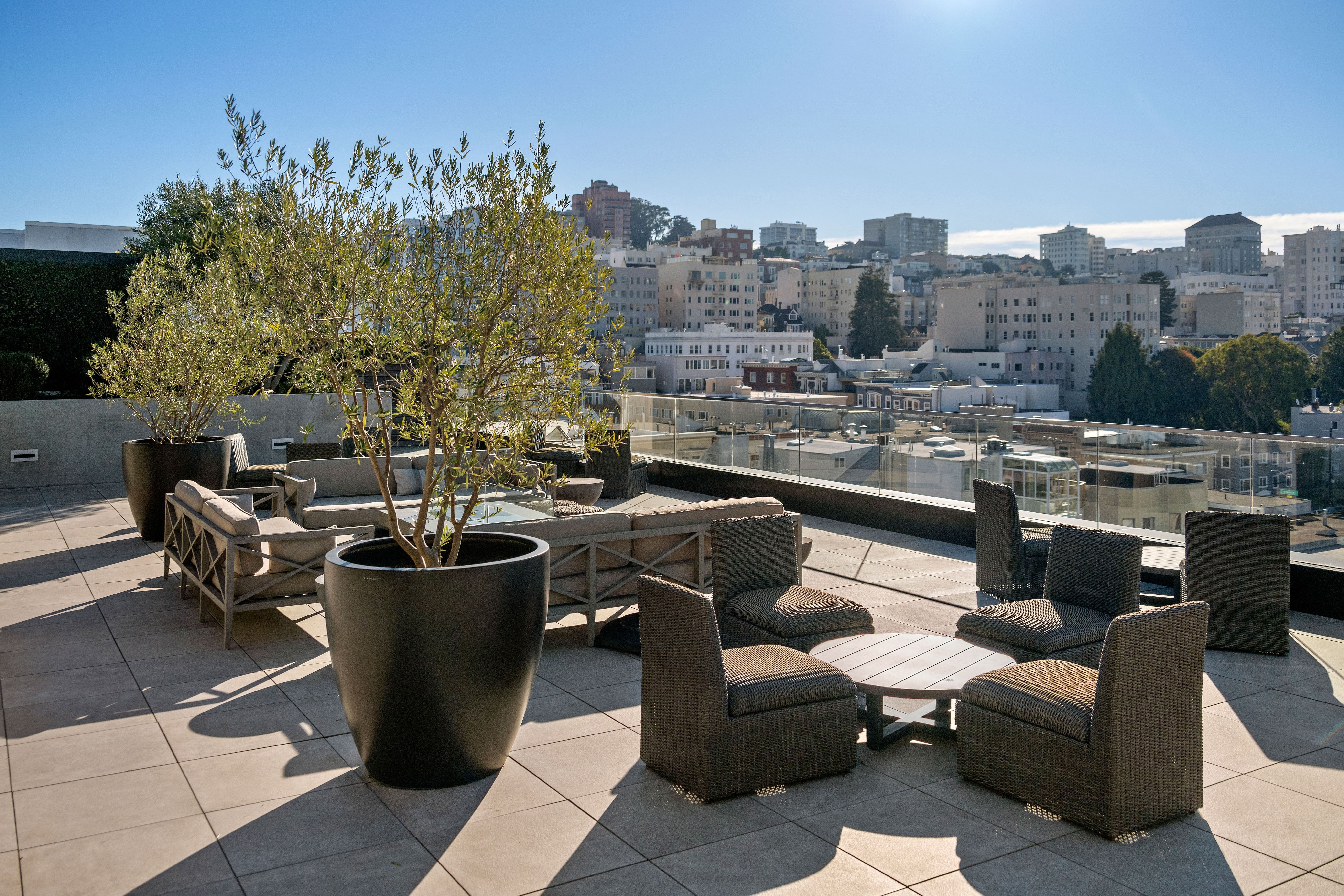 This sleek rooftop terrace offers a sophisticated outdoor living space, complete with modern wicker seating arrangements and potted olive trees. The area is enclosed by a minimalist glass railing, providing an unobstructed, sweeping panoramic view of the urban city skyline. Its spacious, tiled layout makes it an ideal venue for high-end entertaining and relaxation.