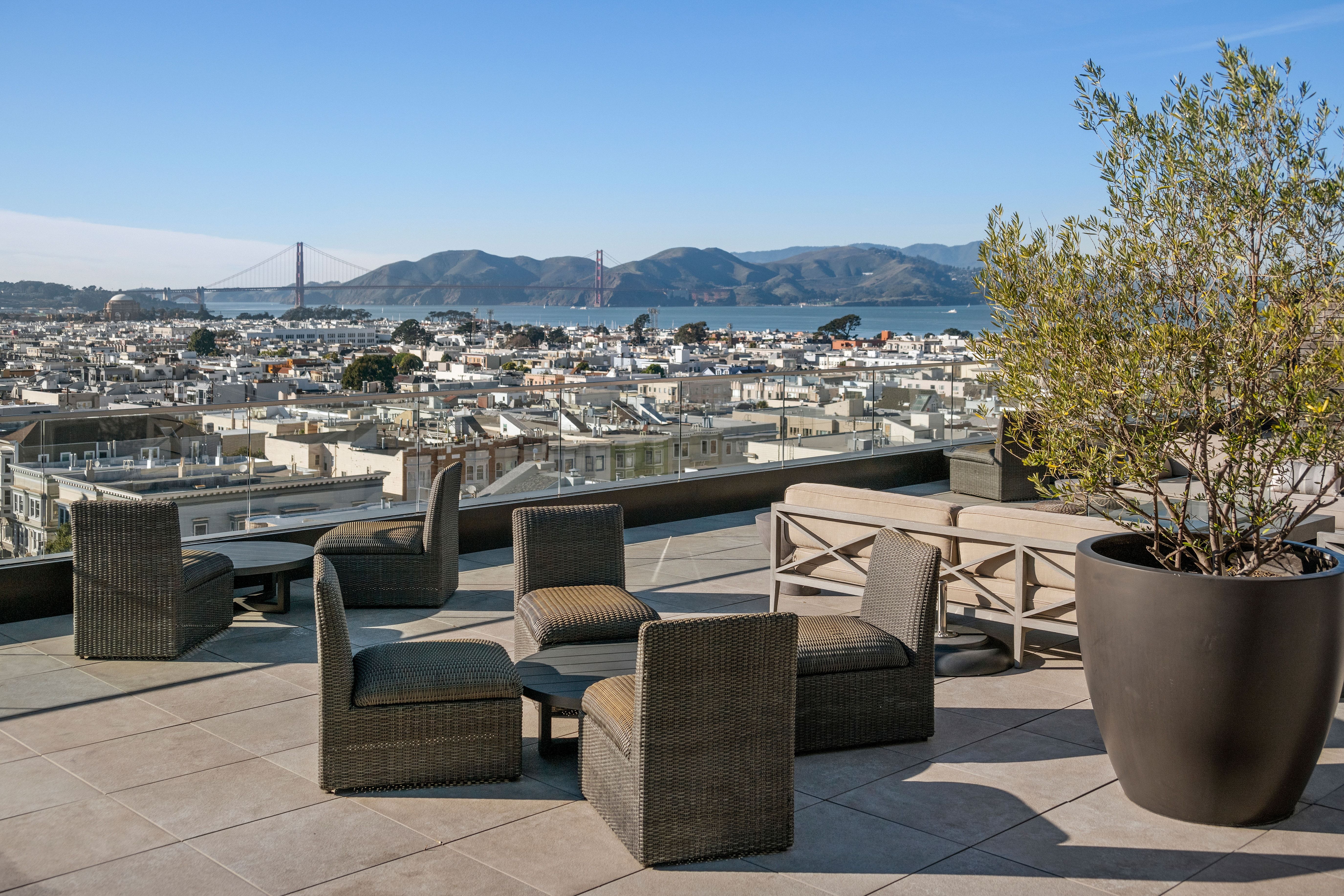 This rooftop terrace offers a stunning panoramic view of San Francisco, including the Golden Gate Bridge and the cityscape. The space features stylish, modern wicker seating arranged around low tables, complemented by an elegant potted olive tree and glass railings. It is an ideal outdoor lounge area that perfectly blends luxury, comfort, and an iconic urban setting.