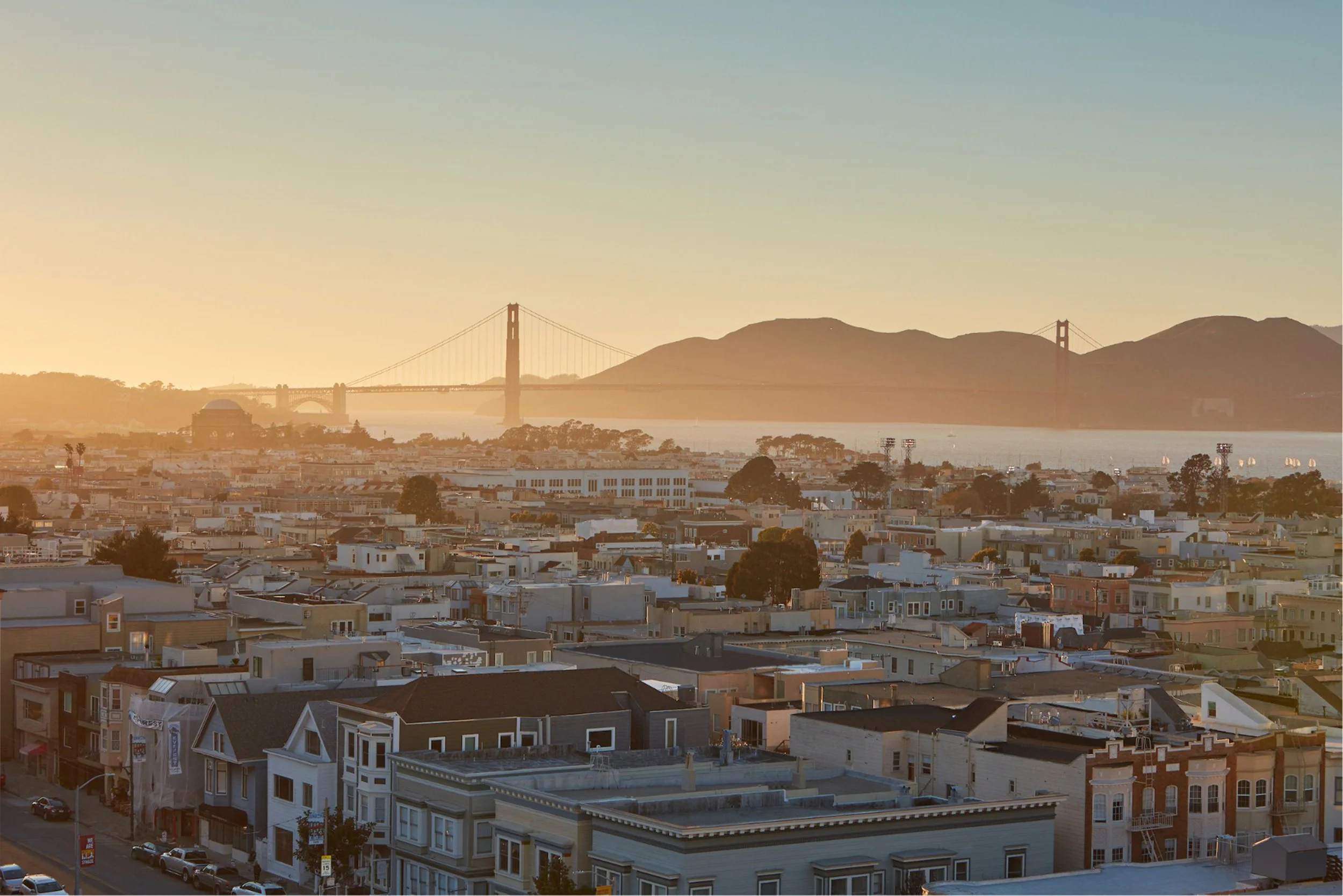 This panoramic aerial view captures a dense urban residential neighborhood in San Francisco, characterized by classic row houses and Victorian-style architecture. The scene is illuminated by a warm, golden sunset light, highlighting the iconic Golden Gate Bridge and the Marin Headlands in the background. It provides an excellent perspective on the city's unique topography, coastal proximity, and the historic charm of the local streetscape.