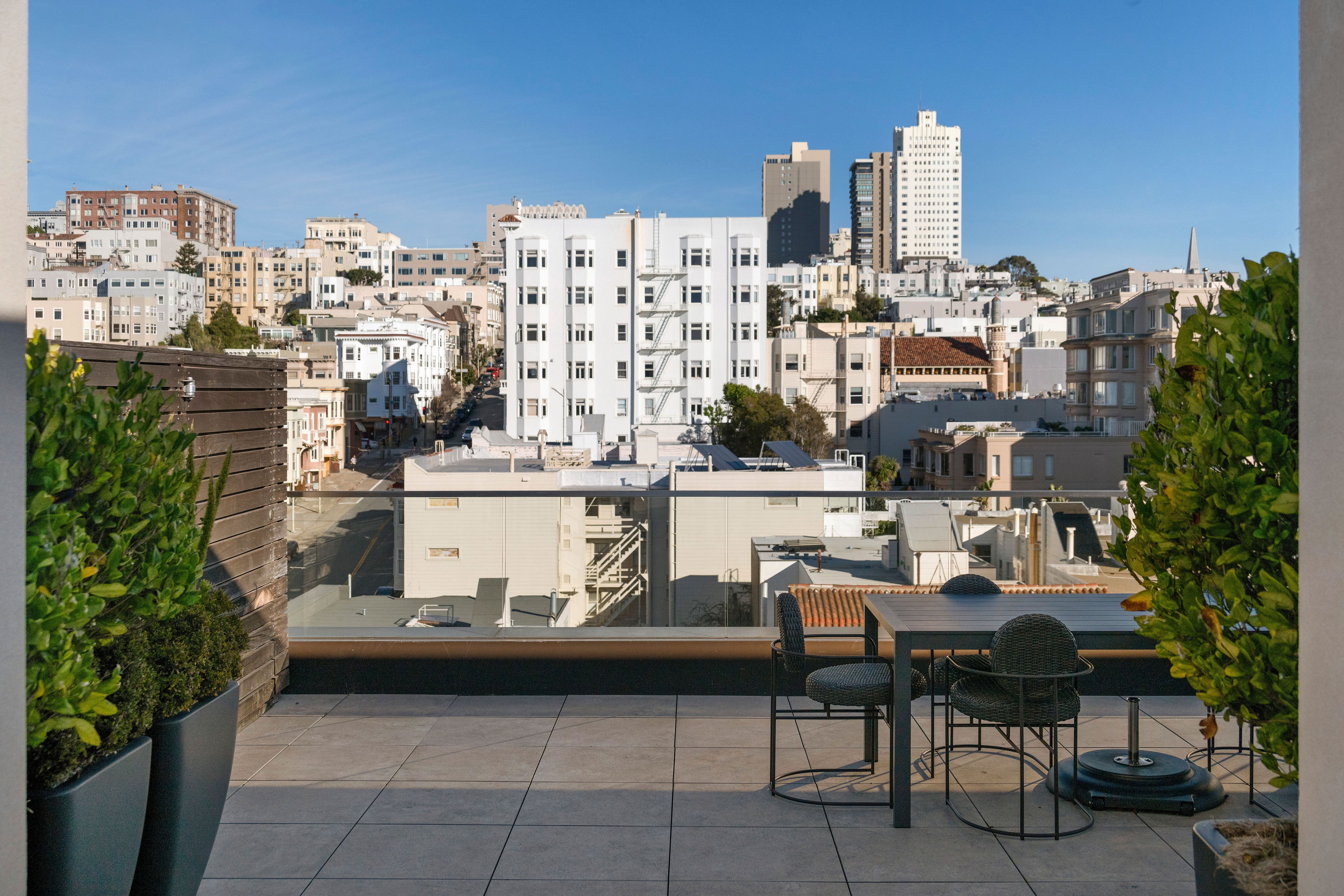 This rooftop deck offers a sophisticated outdoor living space with breathtaking views of the surrounding cityscape. It features integrated planters with lush greenery, a modern table set, and clean, paved tiles, providing a private urban retreat perfect for relaxing or entertaining. The elevated perspective highlights the architectural charm and density of the vibrant neighborhood below.
