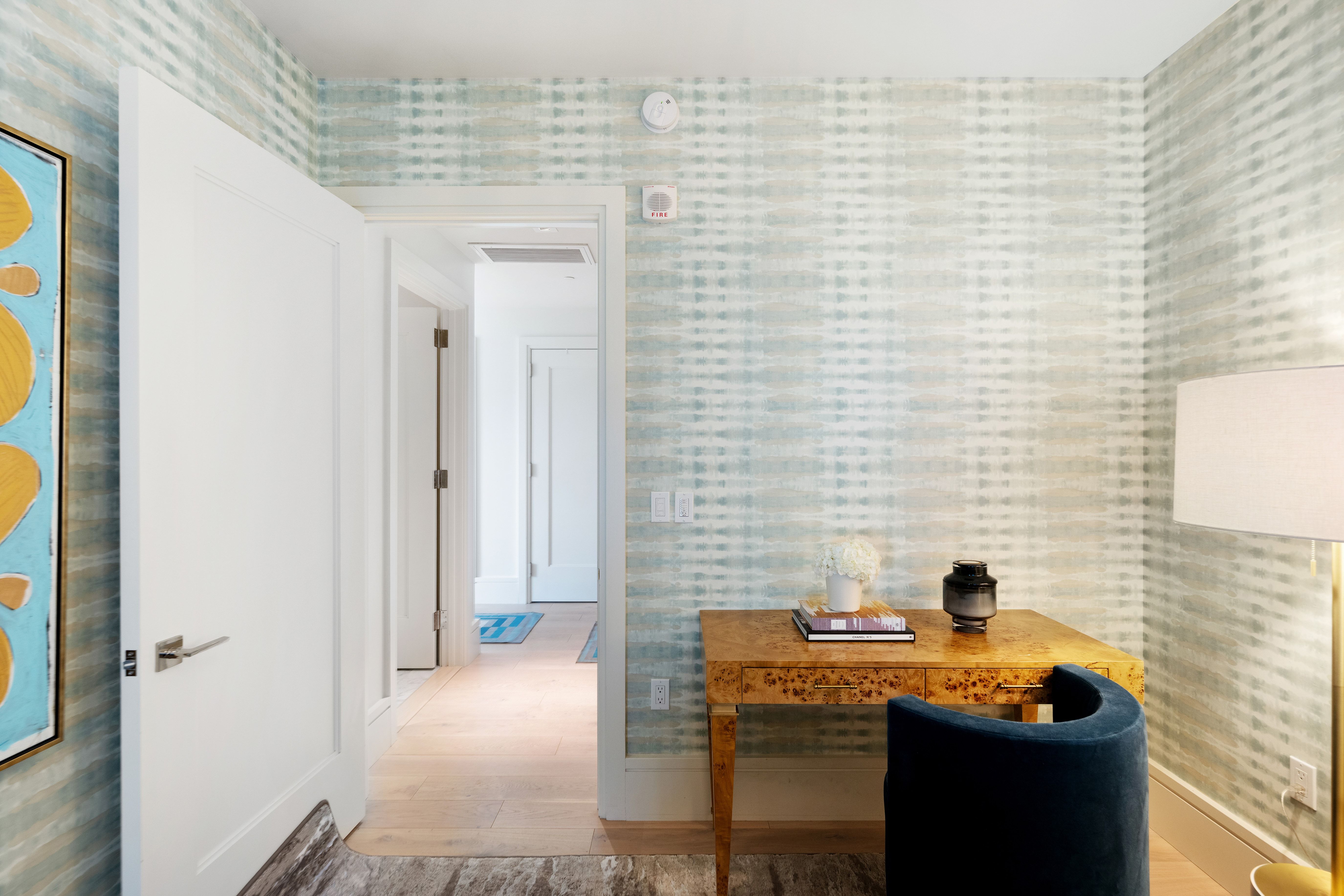 This refined home office or workstation area features a beautiful burl wood desk complemented by a modern navy blue upholstered chair. The space is accented by soft, patterned wallpaper in muted blue and cream tones, creating a calm and professional atmosphere. A doorway leads into a bright, well-lit hallway, highlighting the home's open and thoughtful layout.