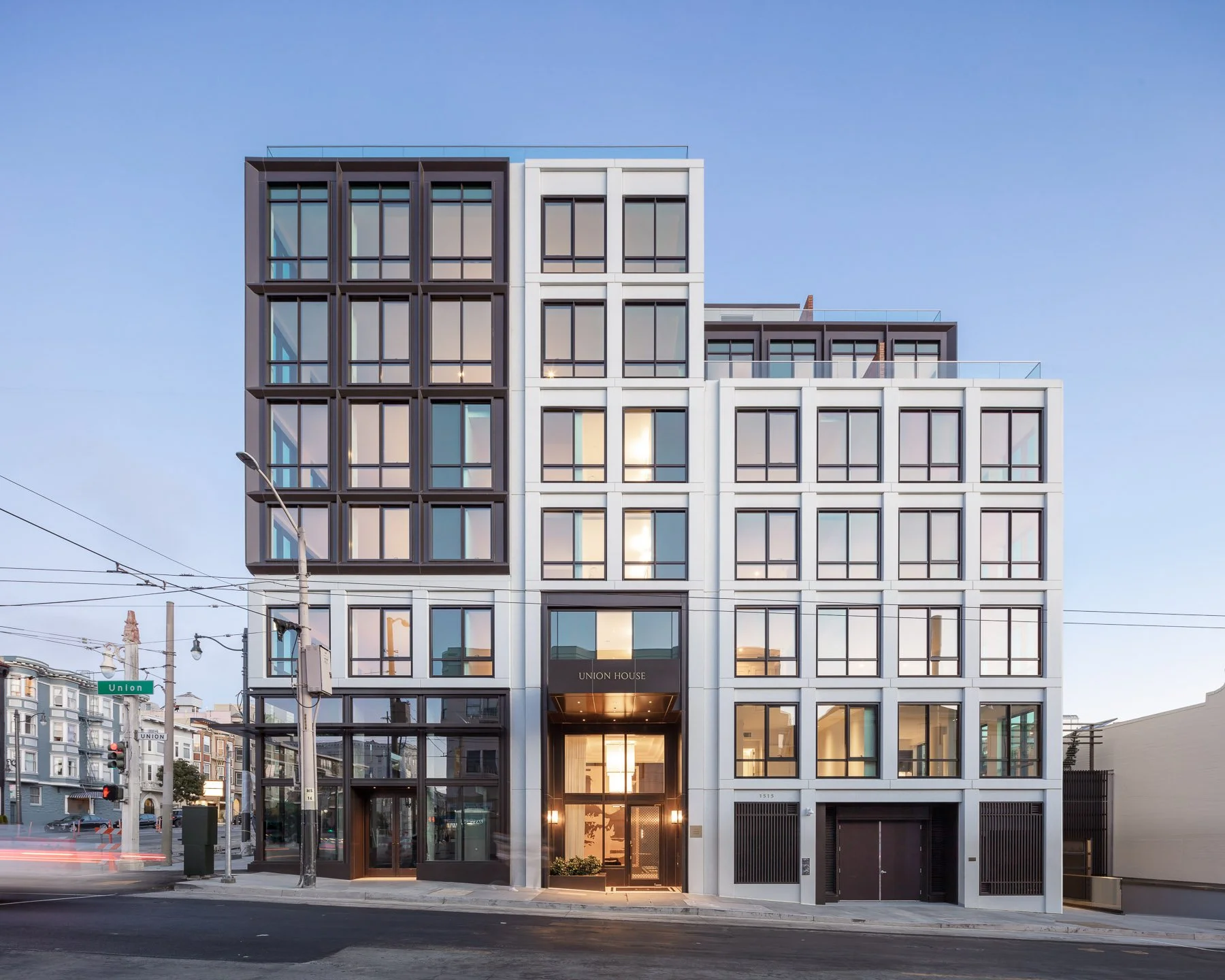 This is a striking, modern multi-story residential building featuring a clean, grid-like architectural facade with a mix of dark charcoal and crisp white finishes. The design incorporates expansive windows that maximize natural light, and the street-level entry is marked by a sophisticated, recessed glass-encased lobby branded as 'UNION HOUSE'. The structure presents a contemporary urban aesthetic ideal for upscale city living.