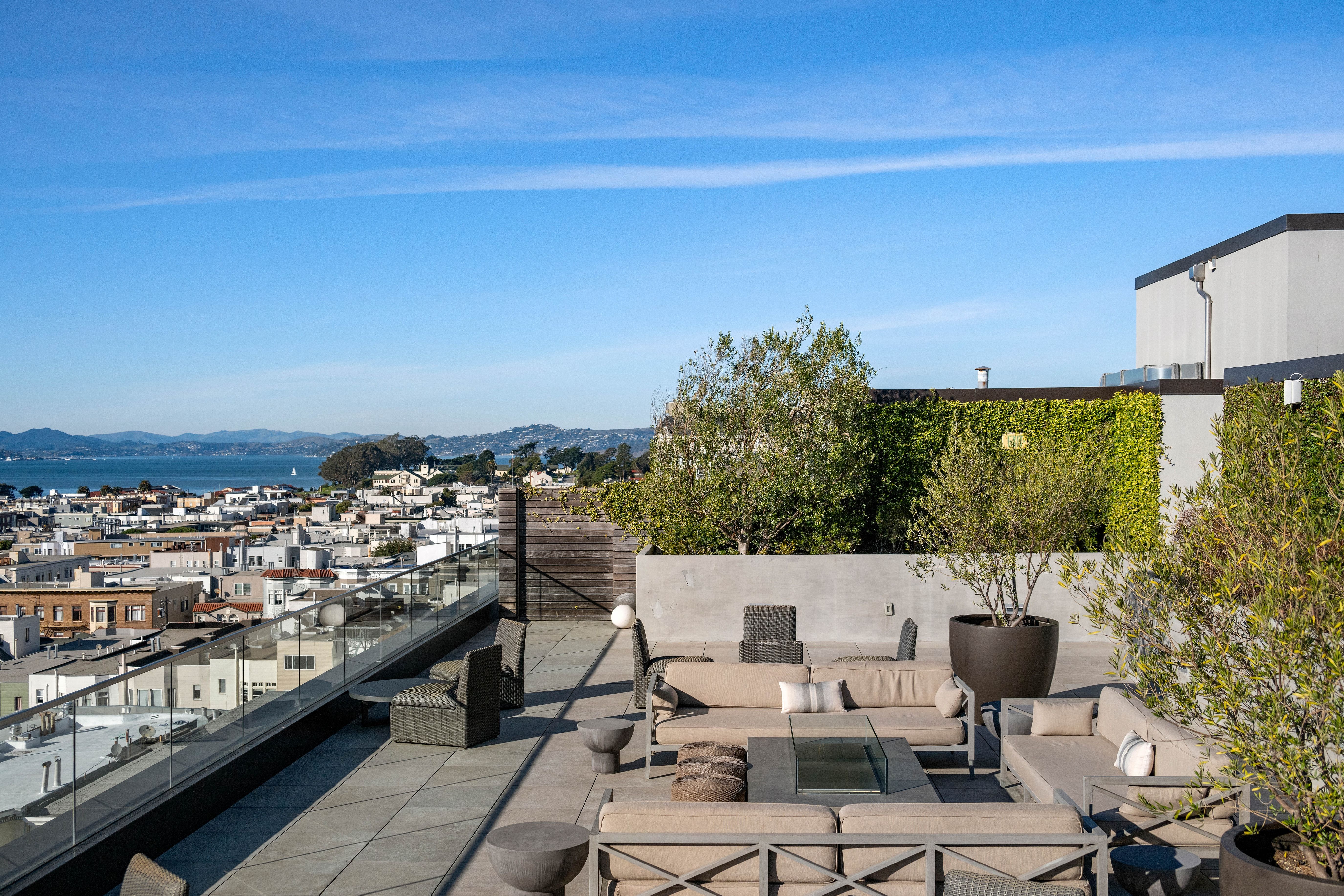 This spacious, modern rooftop terrace offers a sophisticated lounge area with plush sectionals and a central fire pit. Featuring expansive views of the city skyline and bay, the deck is thoughtfully landscaped with potted trees and a green living wall, making it an ideal outdoor entertaining space.