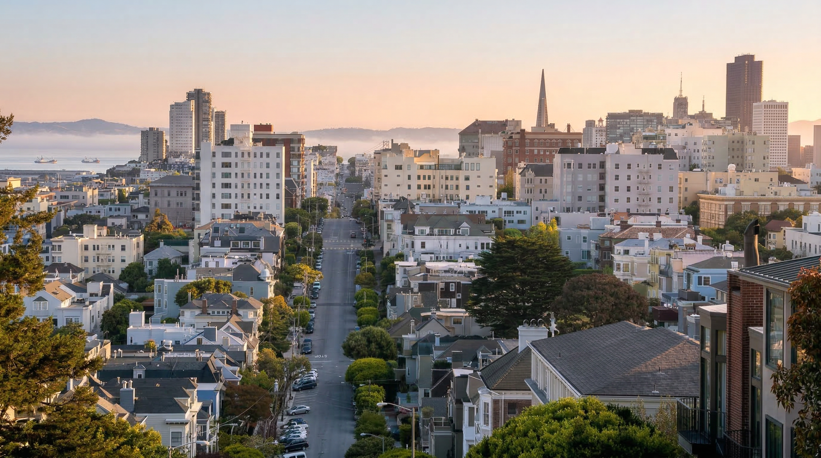 This panoramic aerial view captures a dense urban neighborhood in San Francisco, characterized by historic row houses and multi-story apartment buildings. The street leads the eye toward the distant city skyline, which features iconic structures like the Transamerica Pyramid and high-rise office towers under a soft, golden-hour light. The scene highlights the unique topography and classic architectural density of the city's residential districts.
