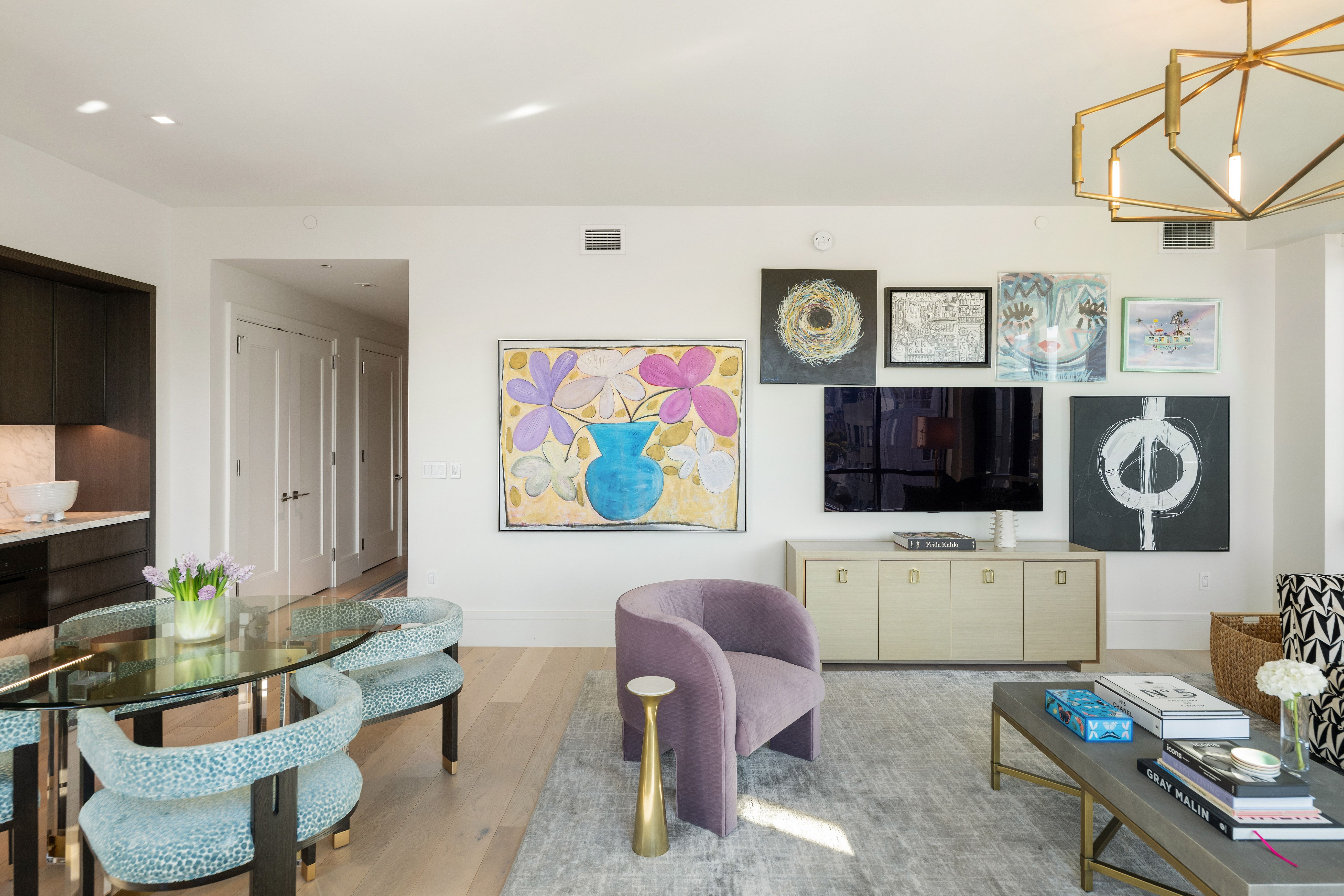 This elegant open-concept living space features a curated gallery wall, a sophisticated lavender velvet accent chair, and a neutral-toned media console. The room blends living and dining areas seamlessly with light hardwood flooring, contemporary designer lighting, and tasteful modern art pieces. The atmosphere is upscale and refined, showcasing a high-end, professionally curated interior design aesthetic.