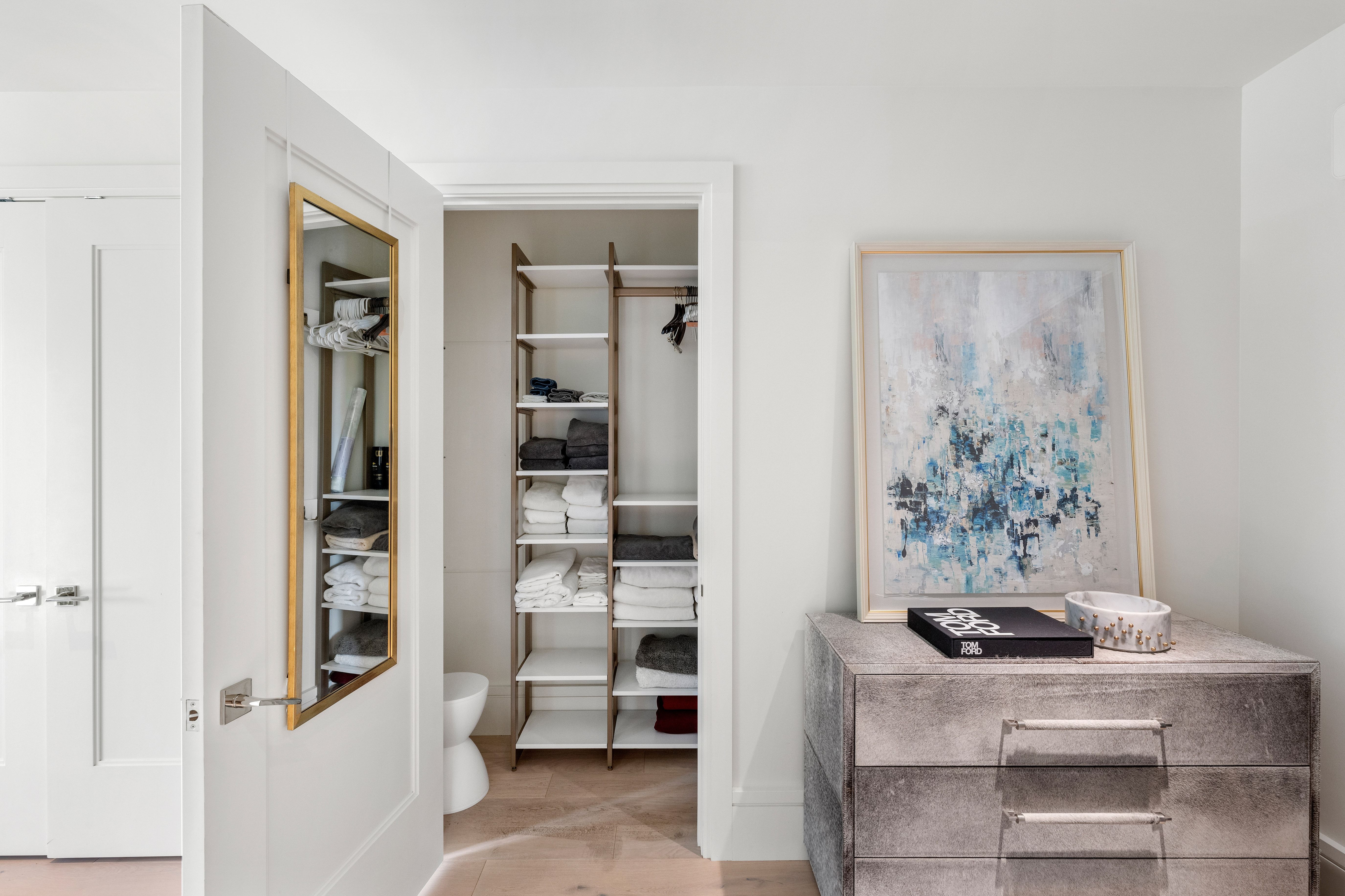 This interior space features a walk-in closet with organized shelving, towels, and clothing storage, providing a clean and tidy aesthetic. Adjacent to the doorway stands a textured, modern chest of drawers topped with a 'Tom Ford' coffee table book and a decorative marble bowl, complemented by contemporary framed artwork. The overall impression is one of sophisticated, organized luxury suitable for a high-end residential listing.