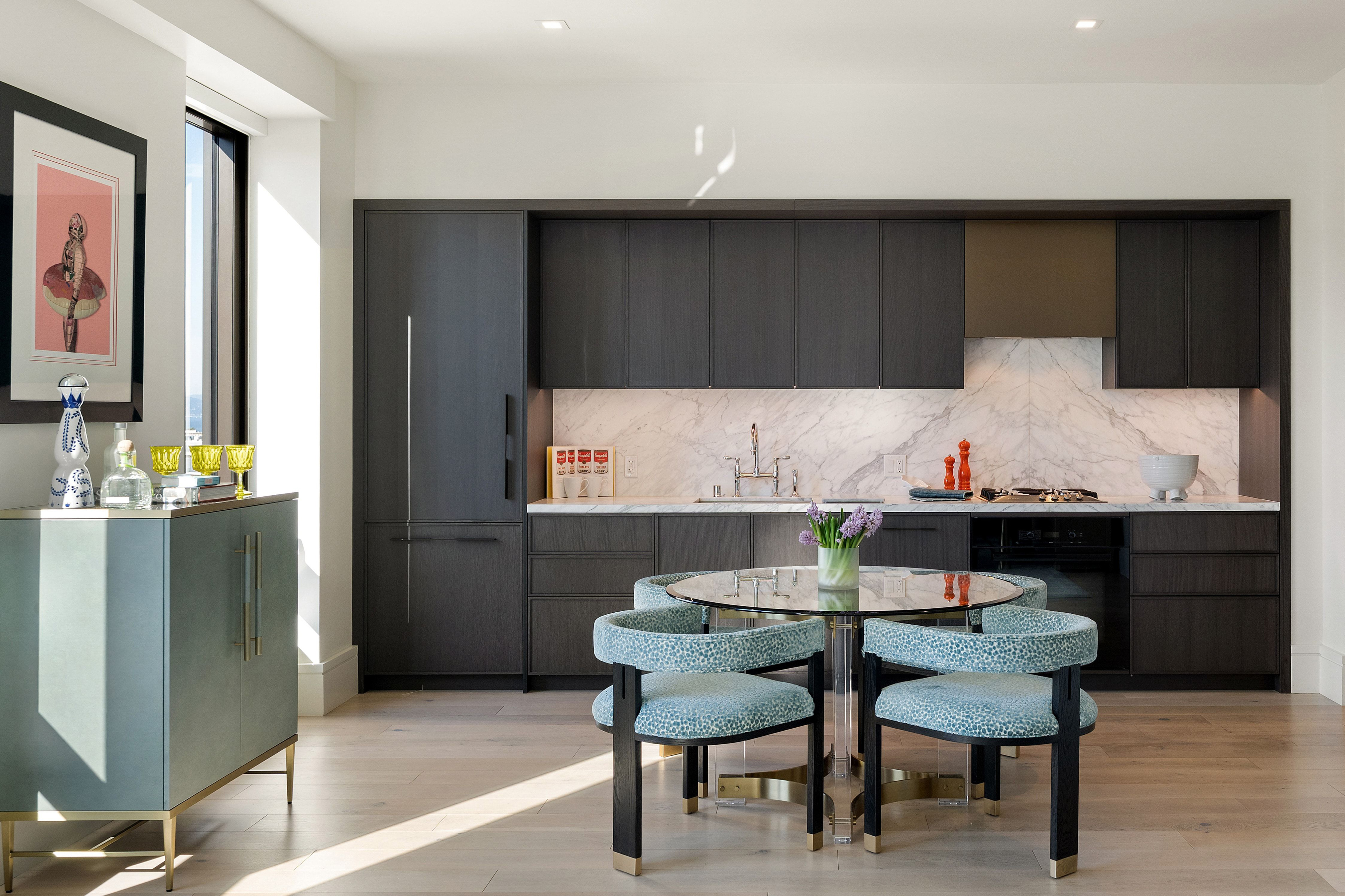 This sophisticated, modern kitchen features sleek dark-toned cabinetry, high-end marble countertops, and a seamless marble backsplash that adds a touch of elegance. A stylish circular dining table with vibrant, patterned blue chairs serves as a central focal point, while light wood flooring and natural light from the side window create an inviting and open atmosphere.